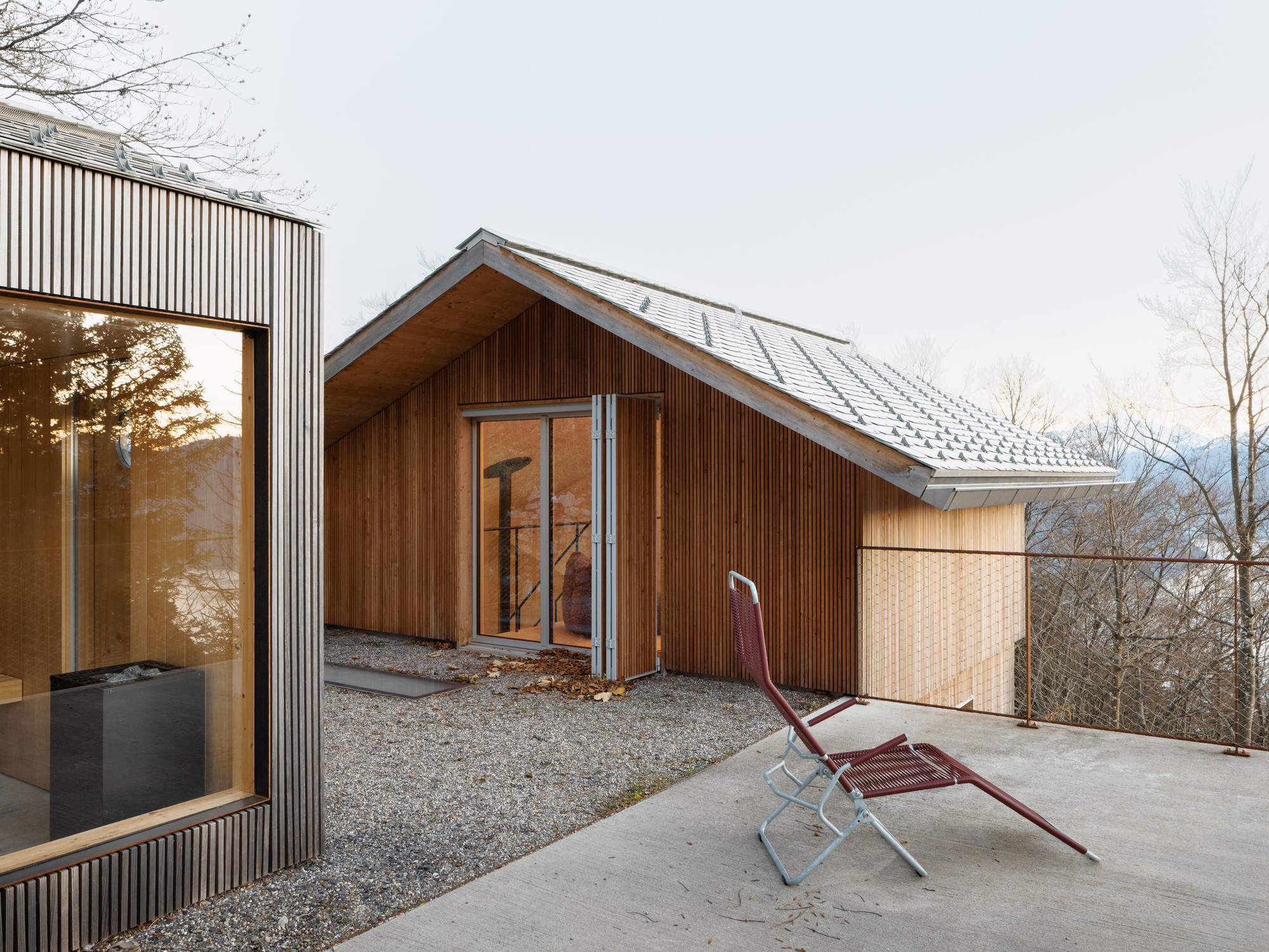 Concrete terrace with a single chair facing timber and metal-clad gabled volumes under overcast skies