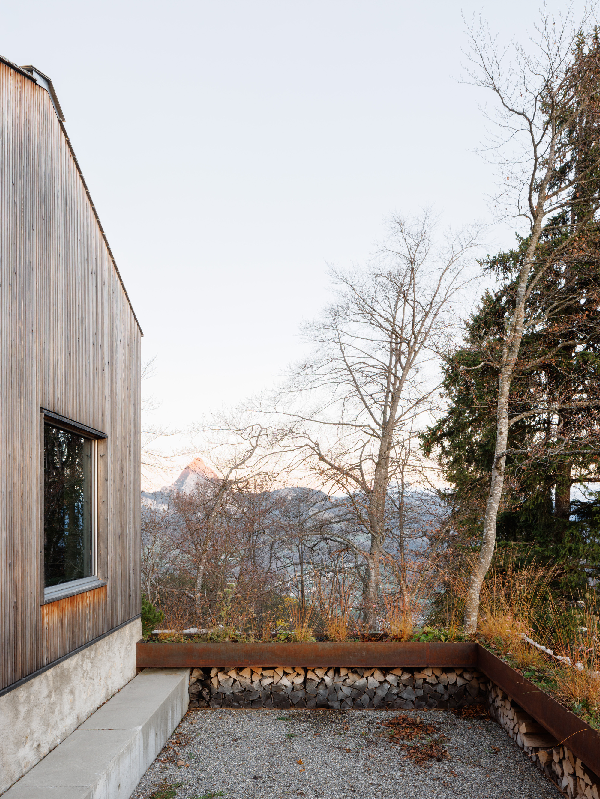 Gravel courtyard with concrete bench and corten steel planter beneath bare trees and distant peaks