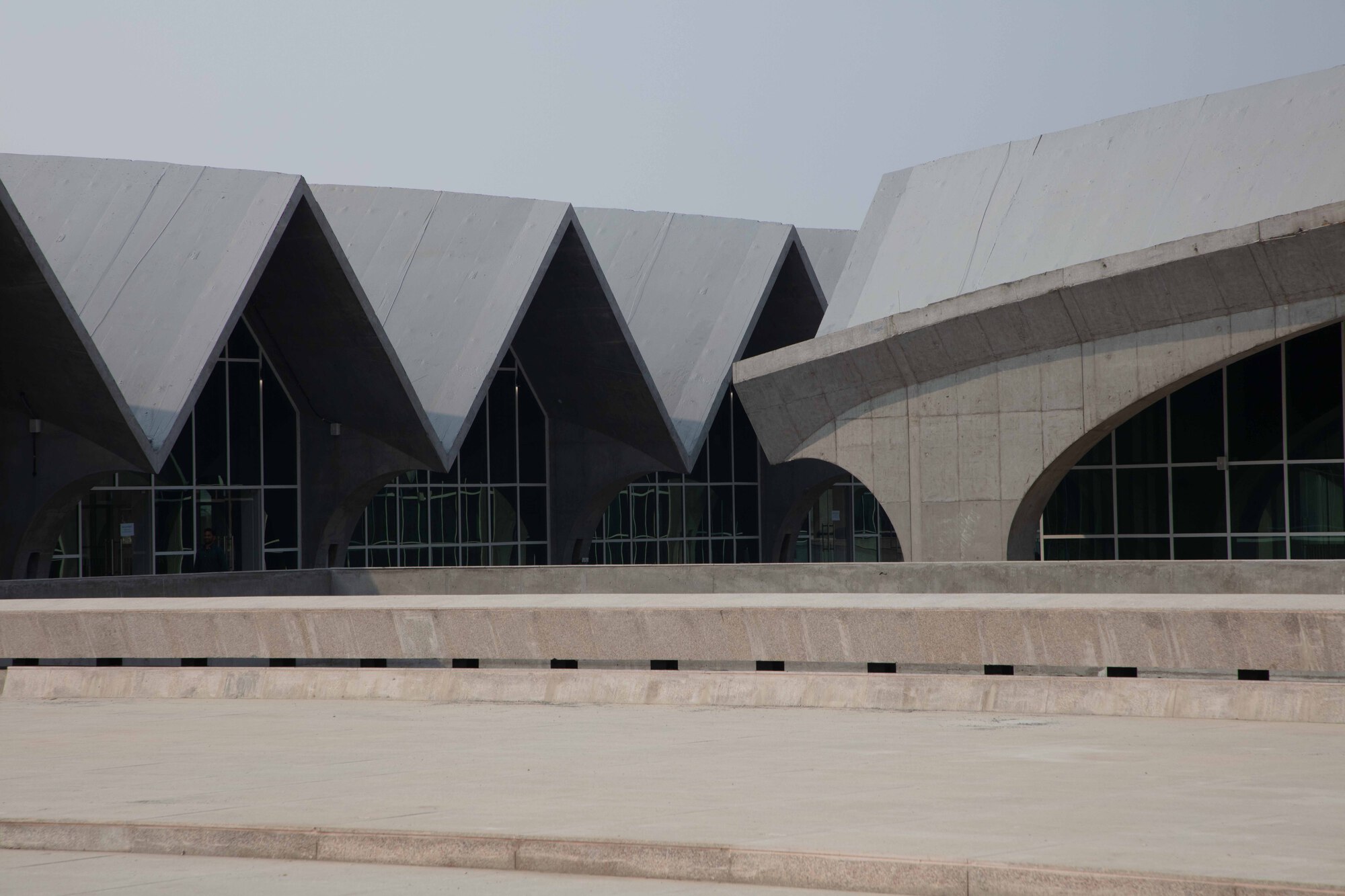 Concrete plaza with stepped seating facing a facade of zigzag gabled roofs and arched glass openings