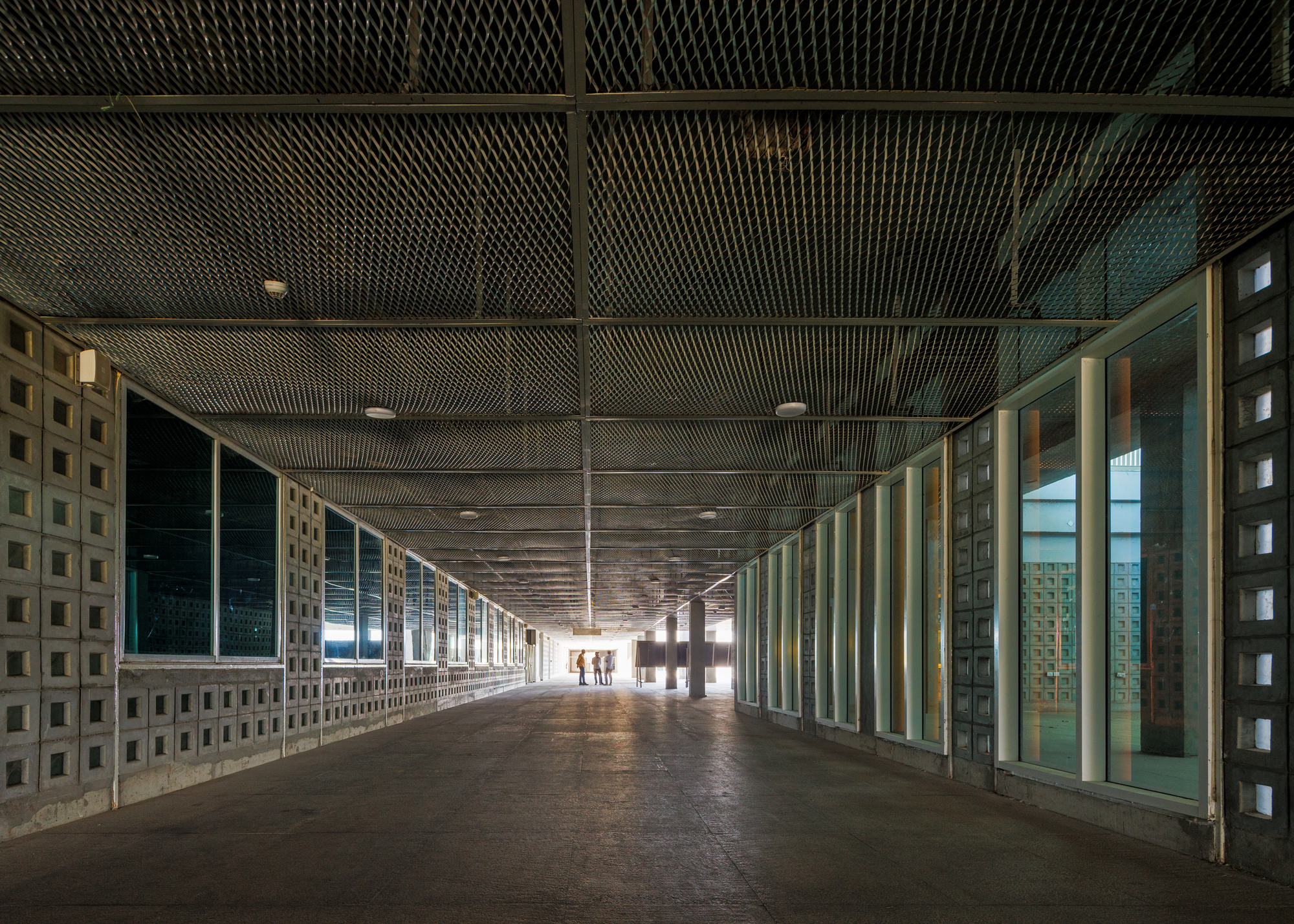 Long corridor with perforated metal ceiling and patterned concrete block walls flanking glazed openings