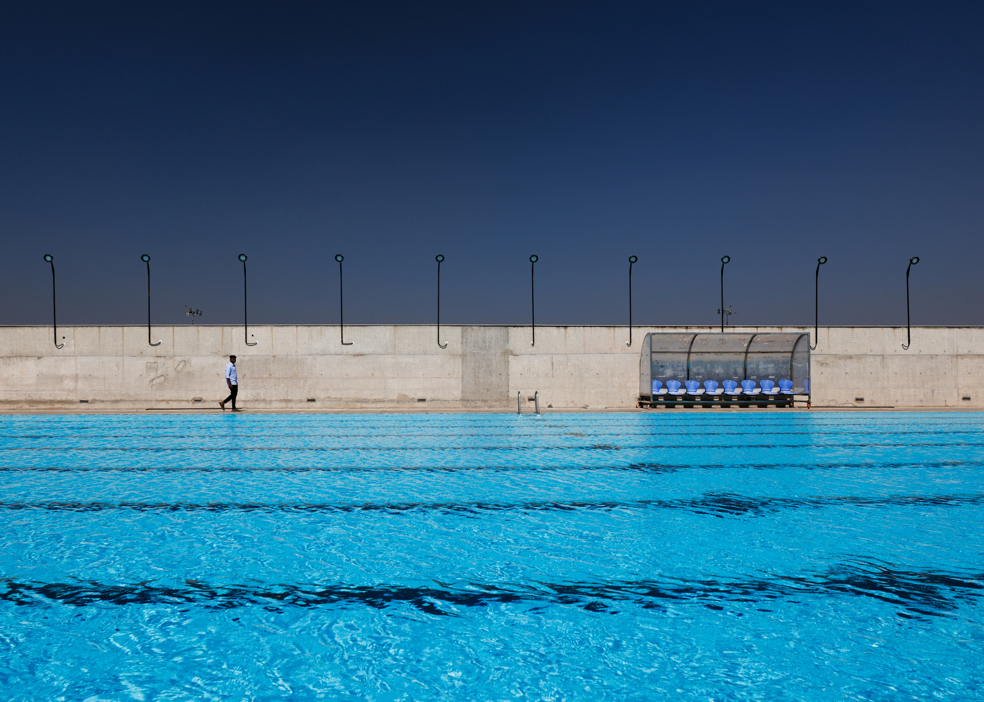 Outdoor pool with lane markers and a person walking along concrete deck with curved metal hooks above
