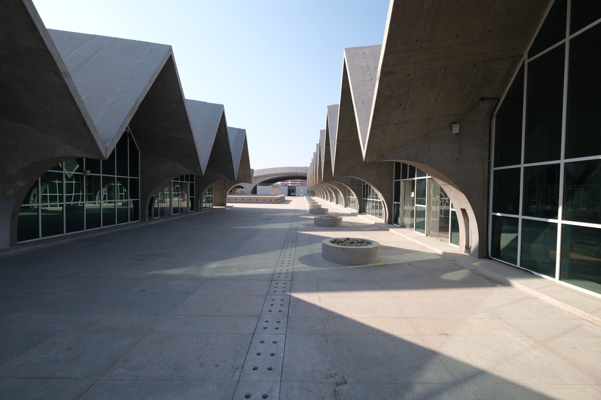Covered walkway between sawtooth roof structures with arched openings and circular planters in morning light