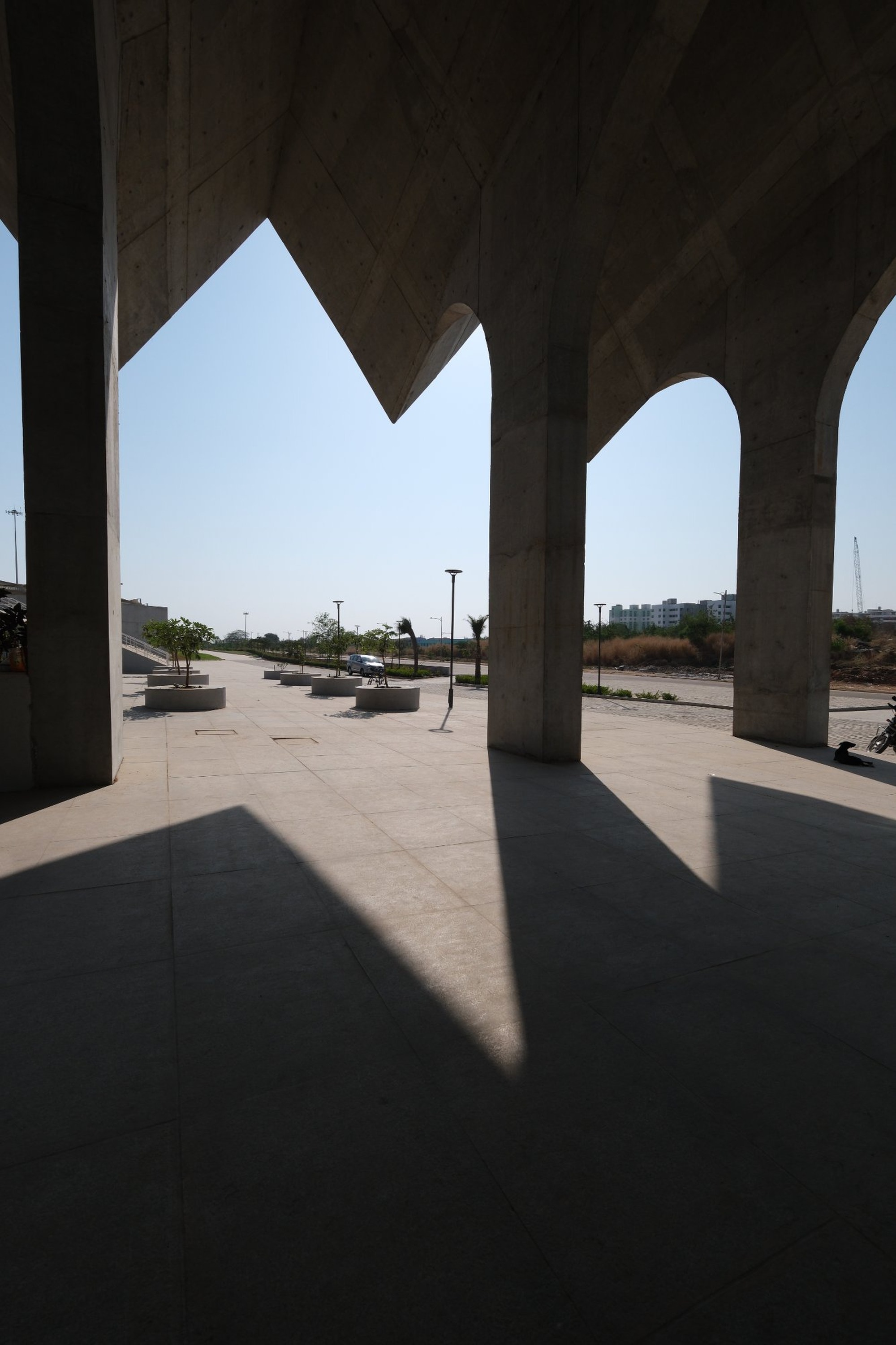 View through angular concrete arches framing a plaza with planters and distant river under clear sky