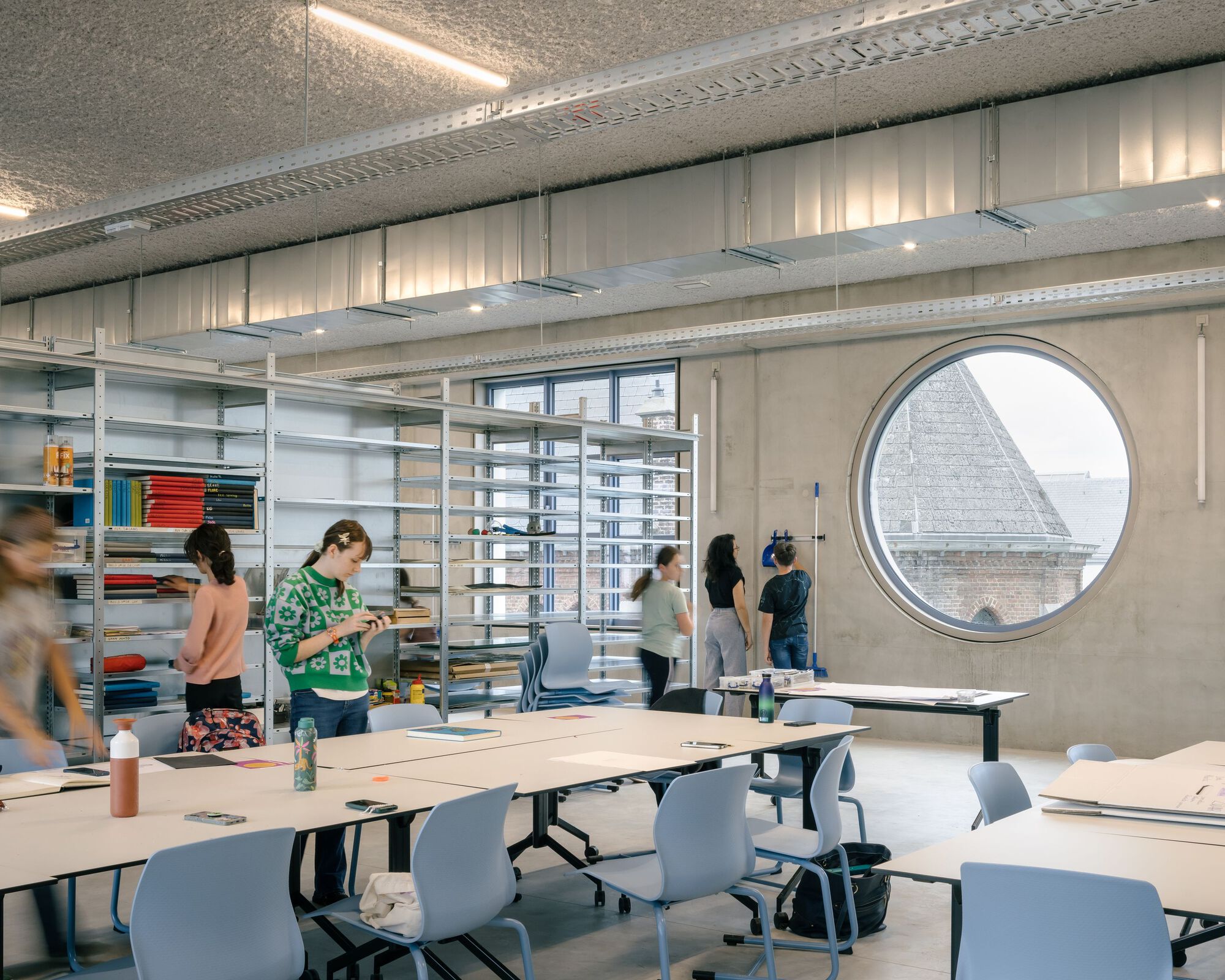 Classroom with exposed concrete ceiling, circular window framing a church roof, and students working at tables