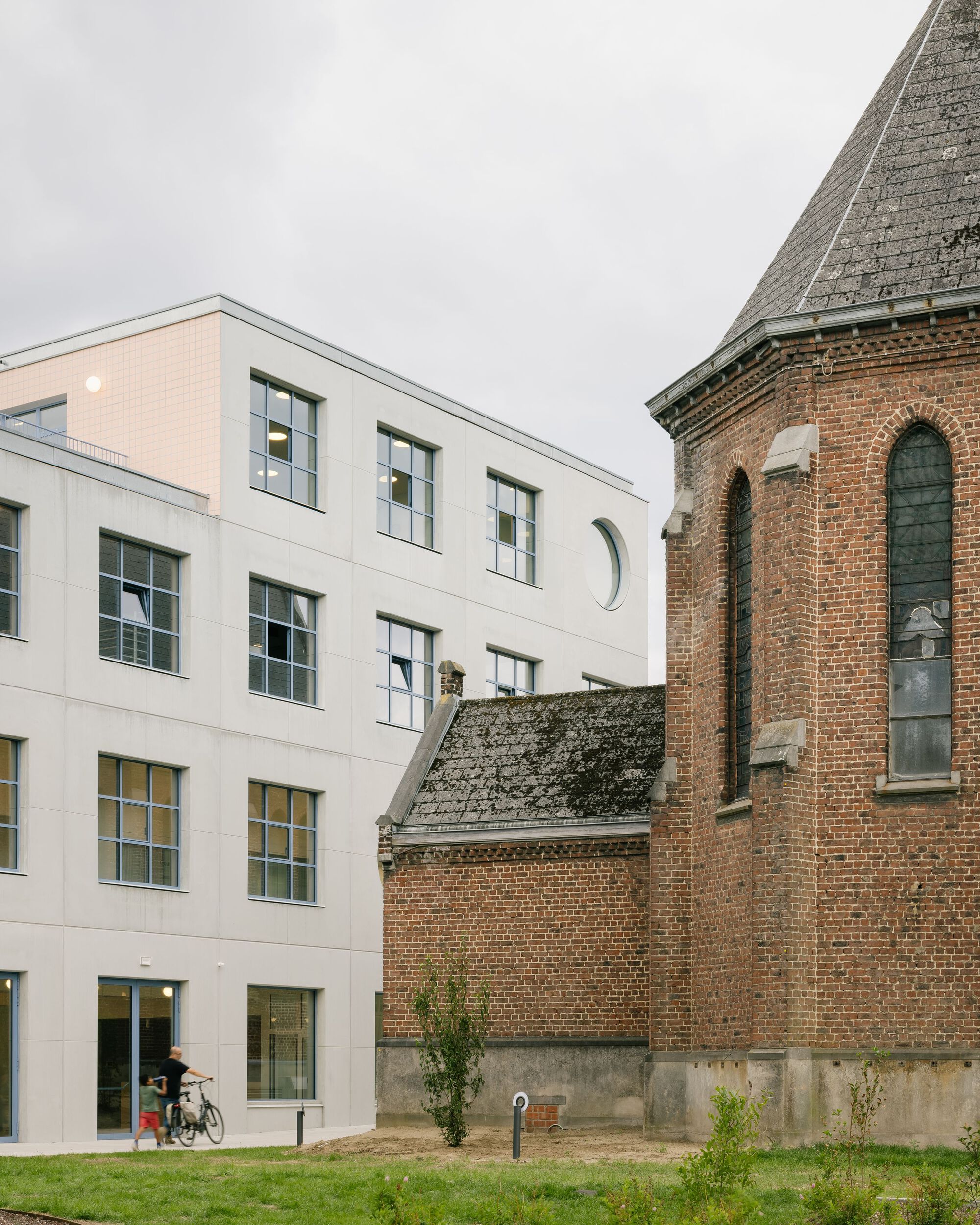White facade with gridded windows and circular portal adjacent to a historic brick church tower