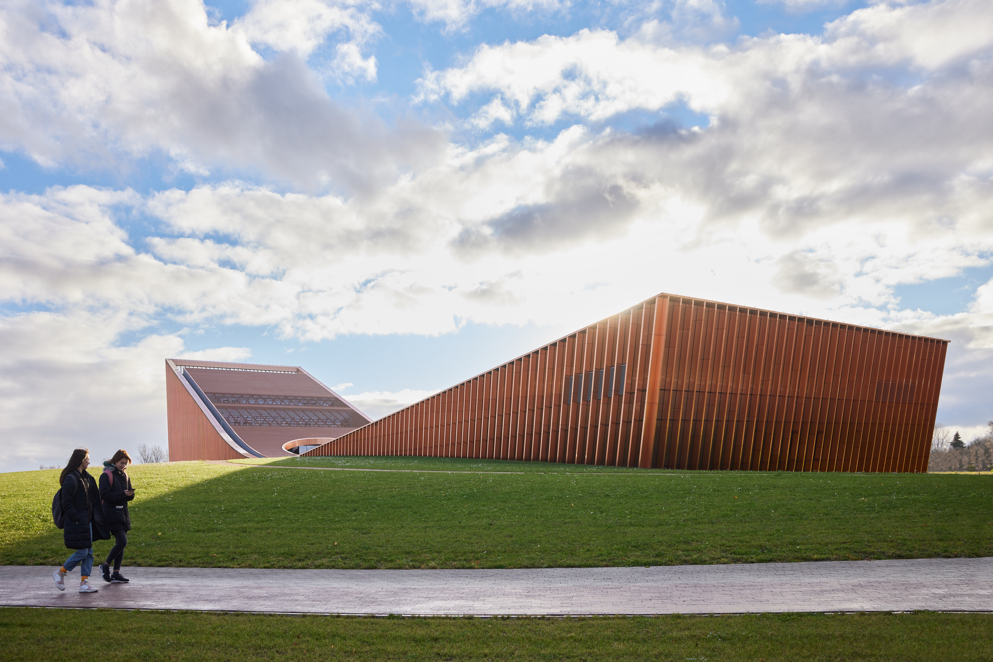 Vertical terracotta-clad facade emerging from a grassed berm with two figures walking along a path
