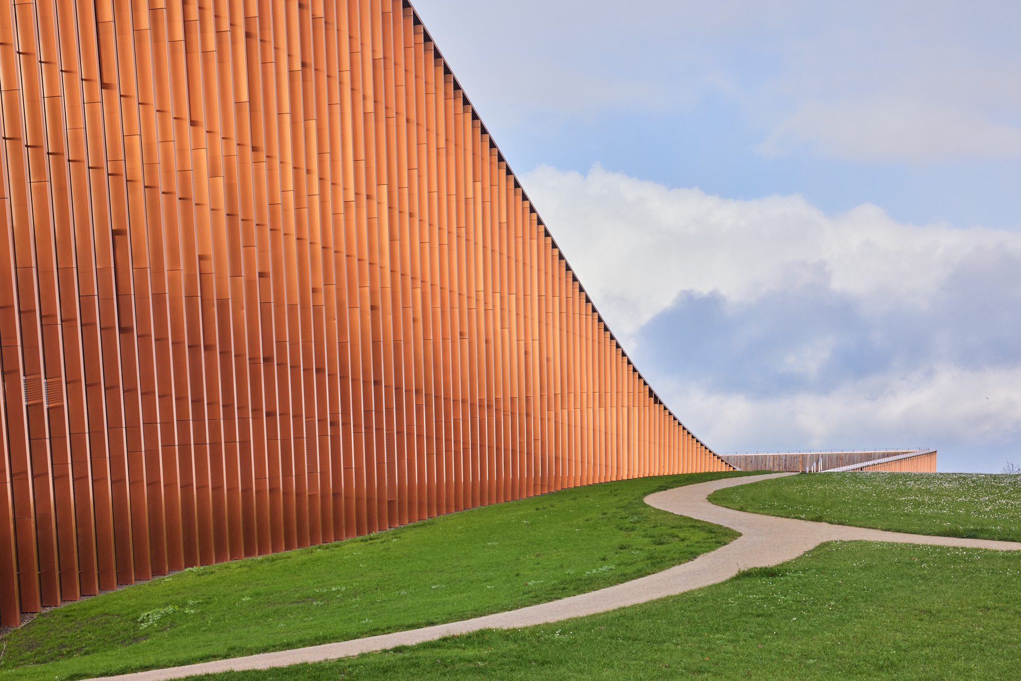 Curved path alongside the vertical terracotta cladding rising out of the grass under cloudy skies