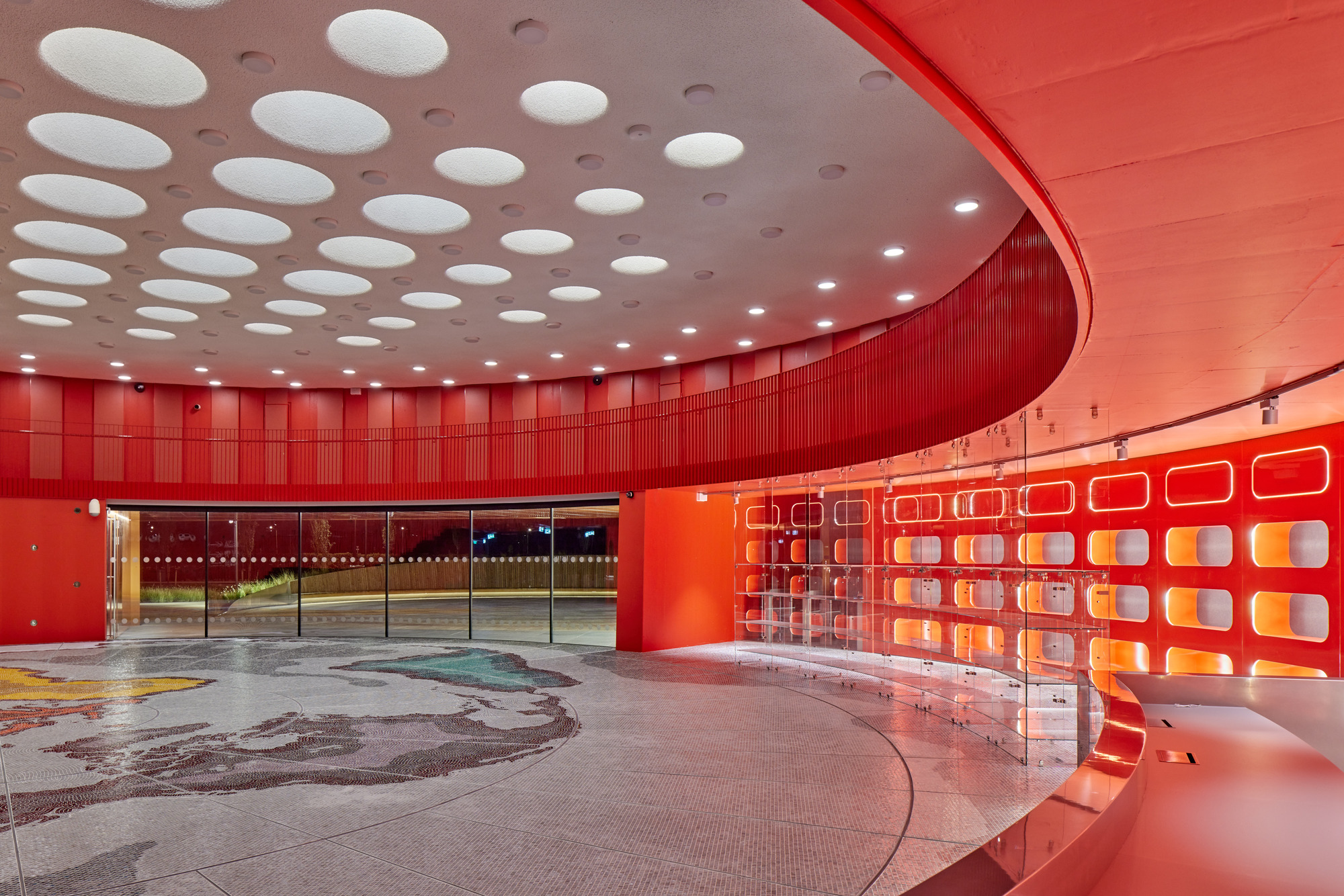 Interior lobby with circular skylights in the ceiling and red illuminated display wall beside a pool