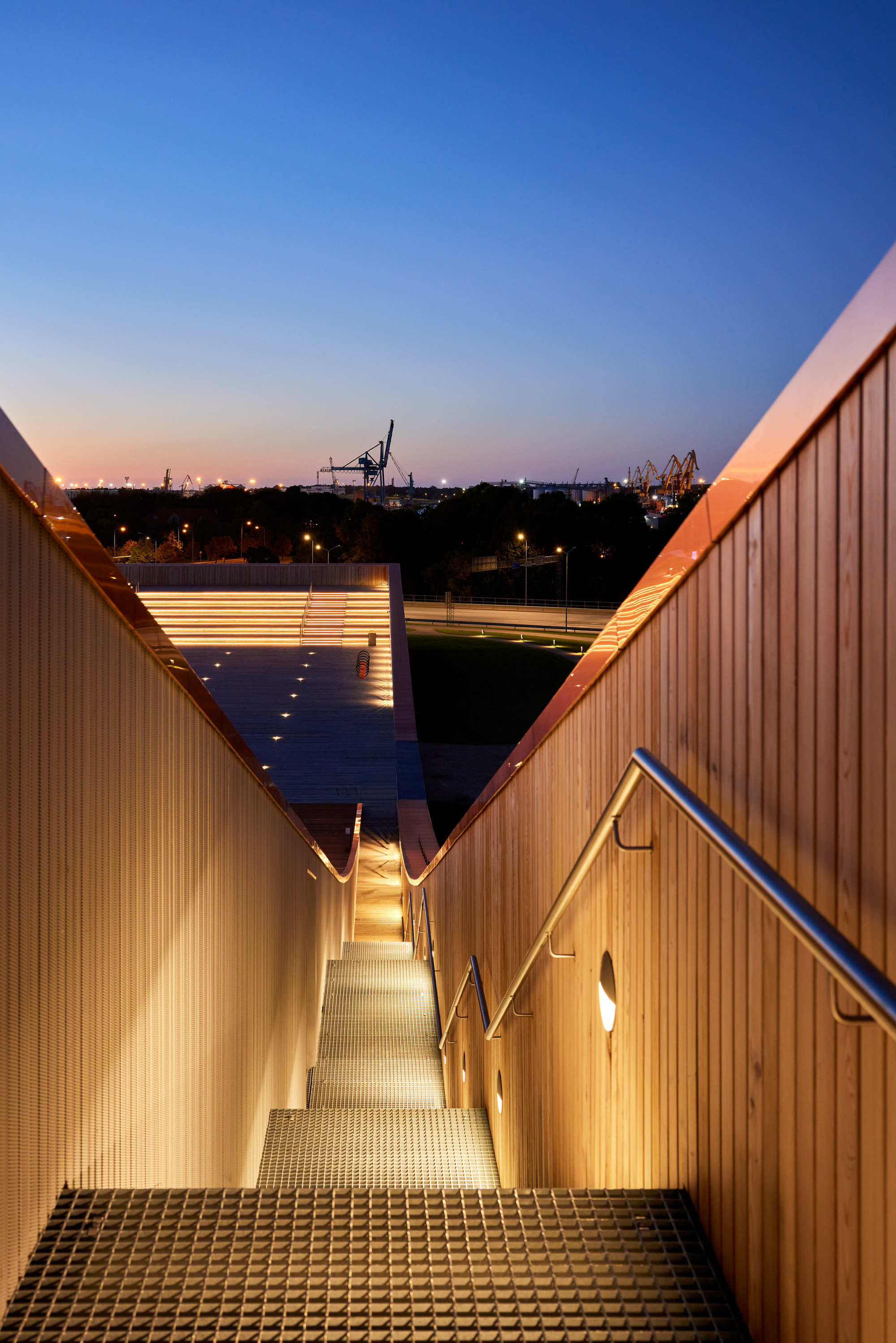 Exterior stairway descending between timber-clad walls with strip lighting at dusk