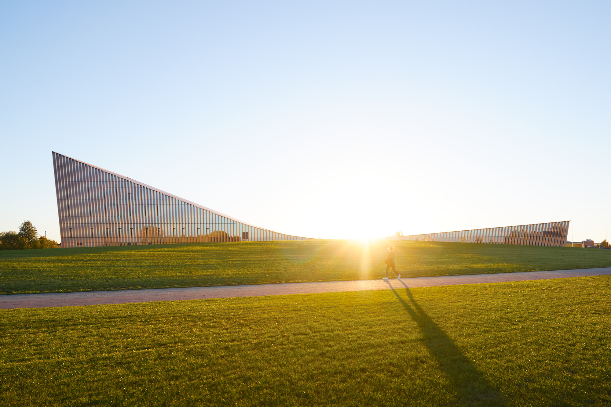 Two angled facade volumes rising from the grass lawn at sunrise with cyclists passing