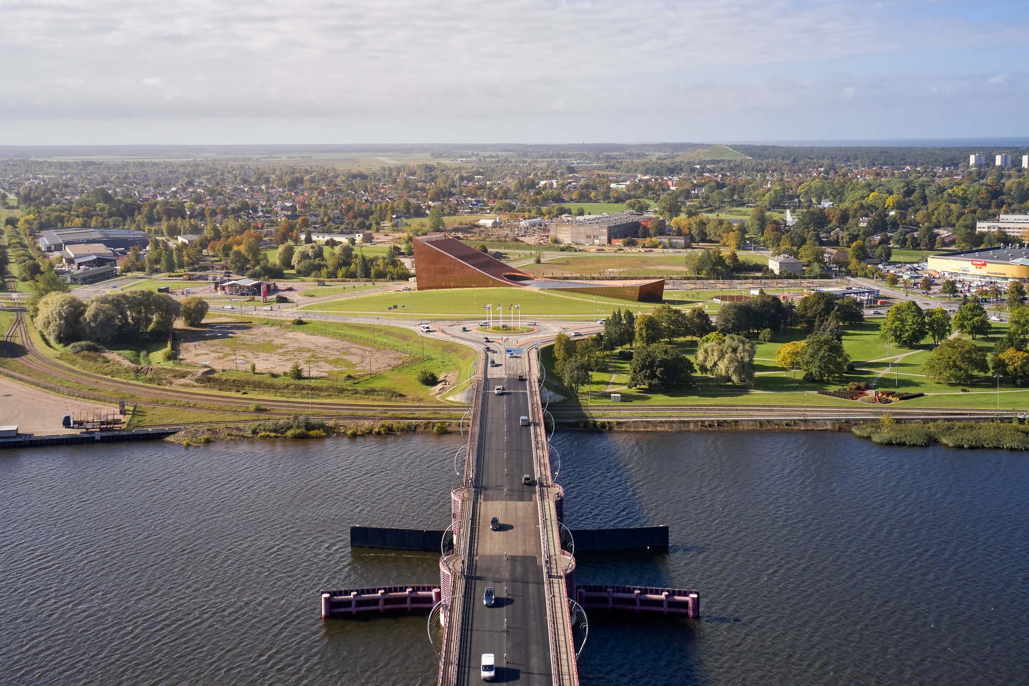 Aerial perspective showing the folded copper roof beyond a pedestrian bridge crossing the river in autumn