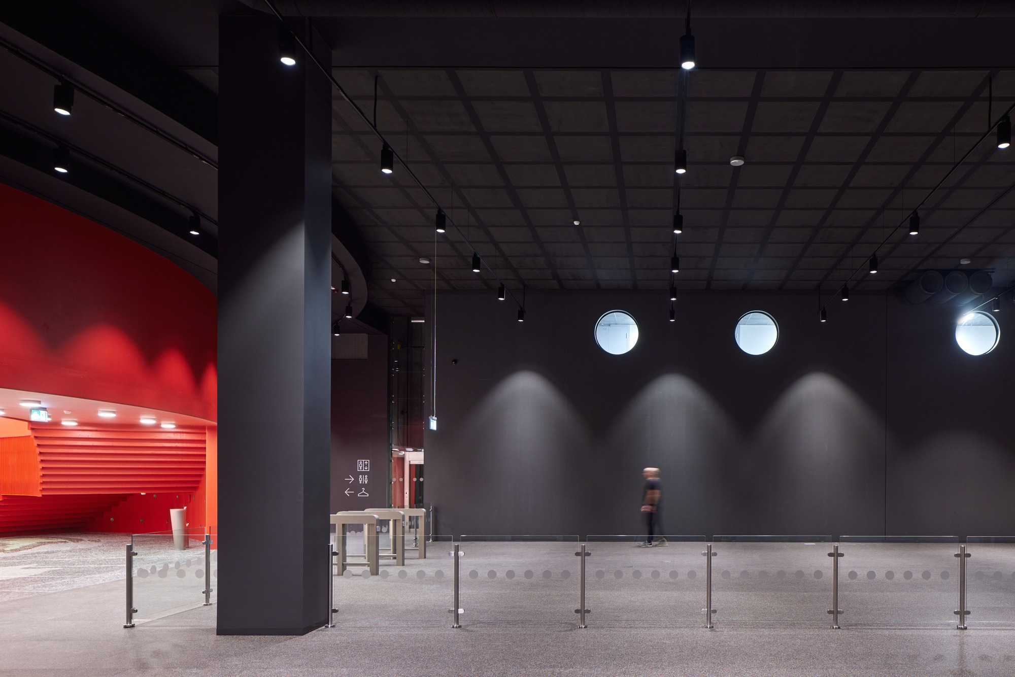 Dark grey interior lobby with circular porthole windows and a figure walking past metal barriers
