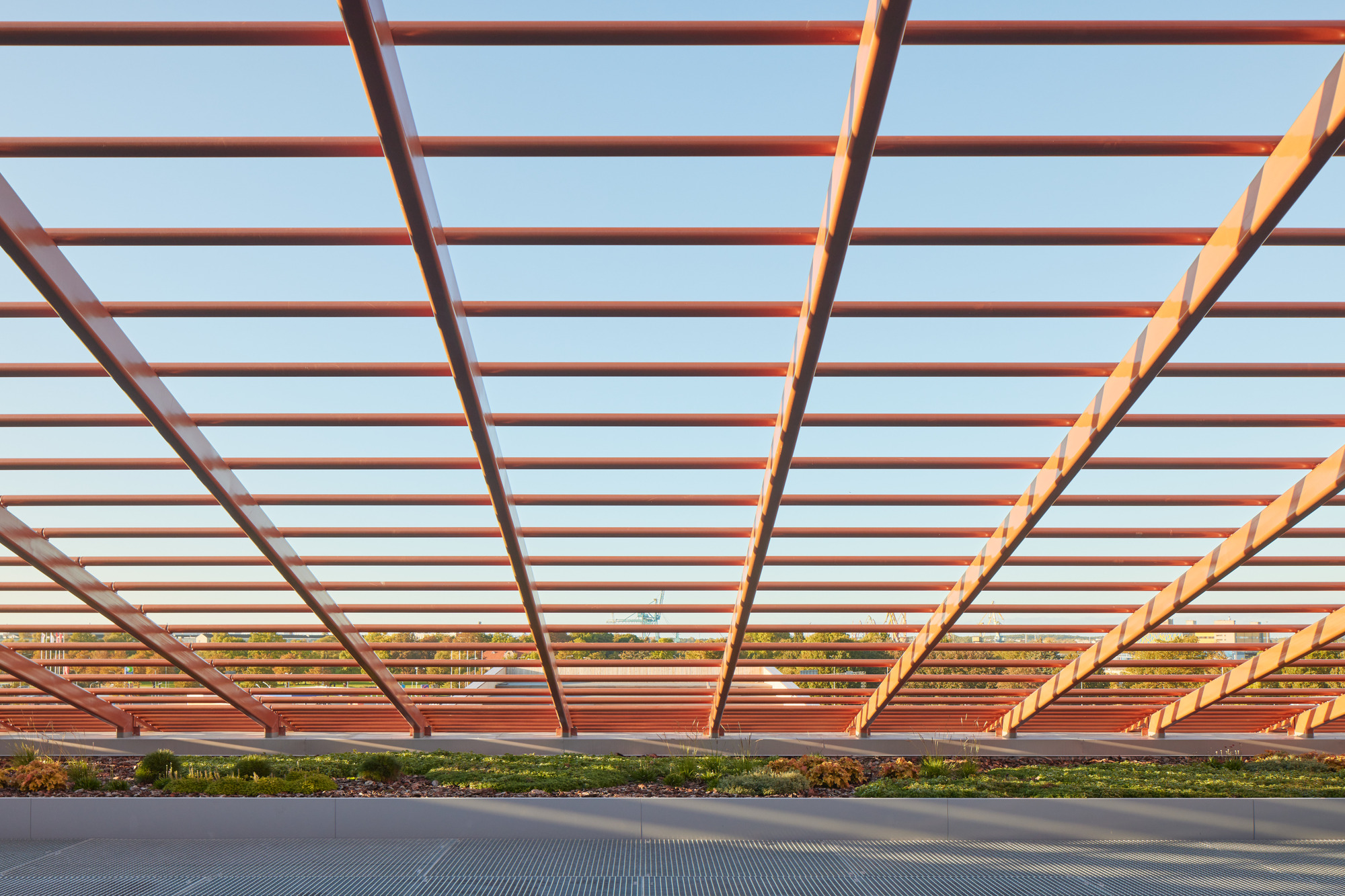 View beneath a pergola with red steel beams and glass infill panels at sunset