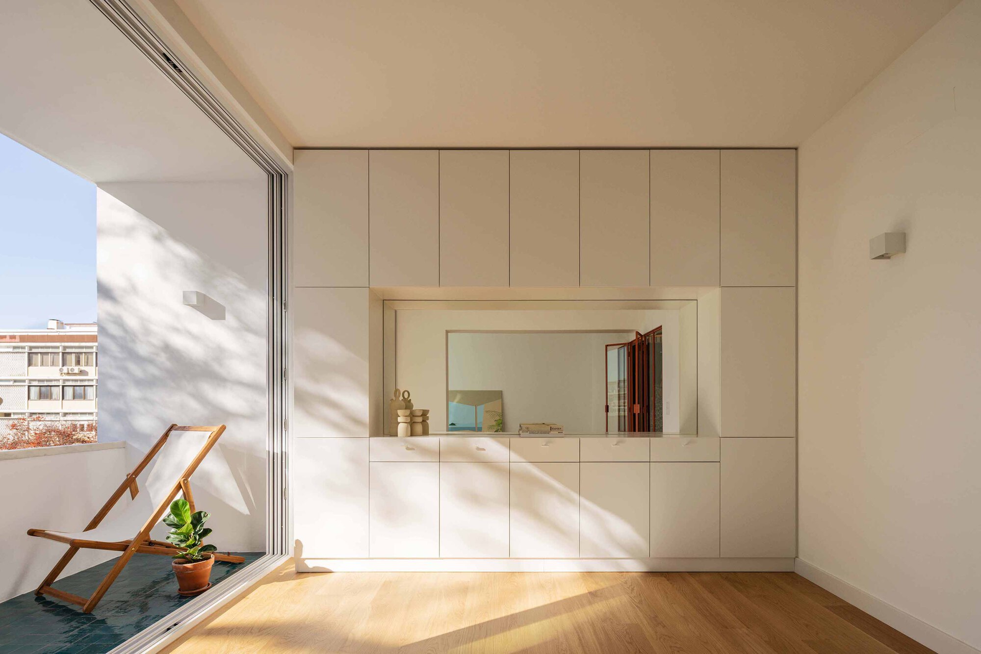 White storage wall with recessed mirror and sliding terrace door opening to a deck chair