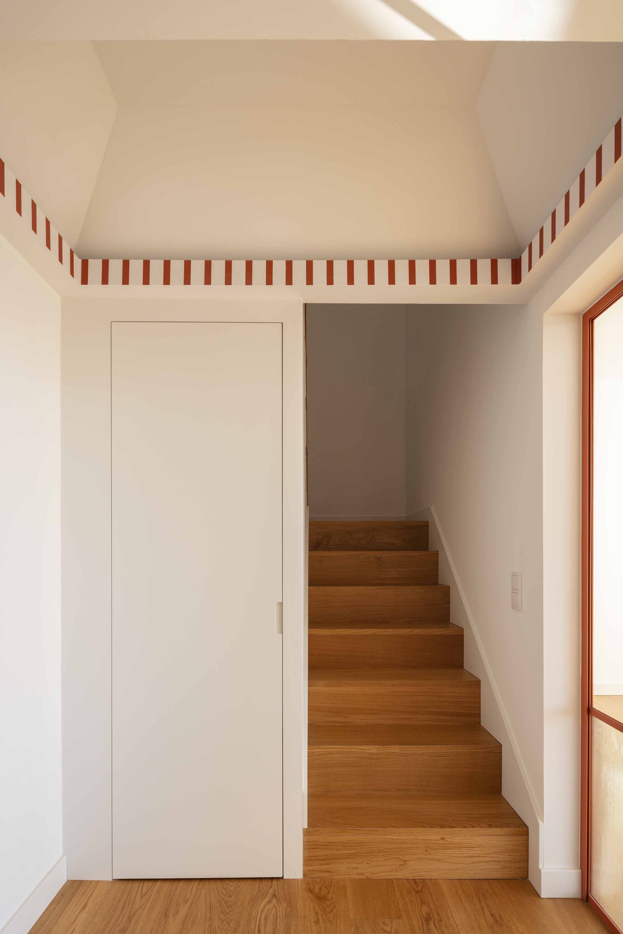 Timber staircase ascending beside a flush white door with red and white striped trim at the ceiling