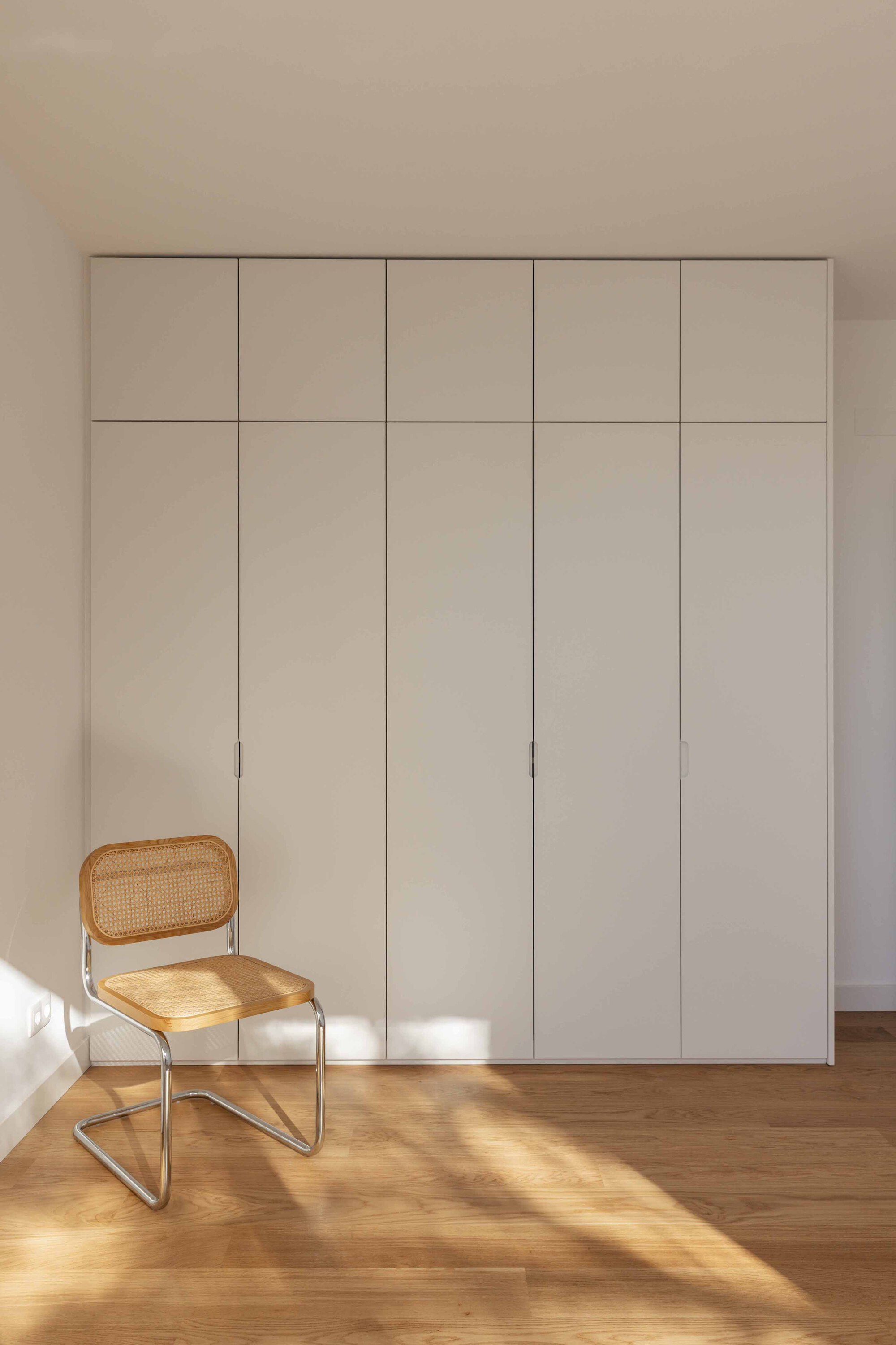 Full-height white storage wall with grid of doors beside a cane-backed chair in sunlight