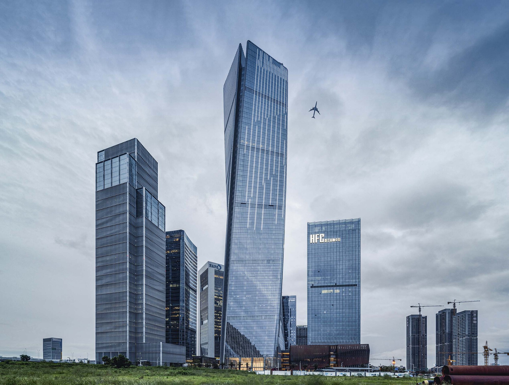 Cluster of glass towers with twisted geometries under a cloudy sky with a plane passing overhead