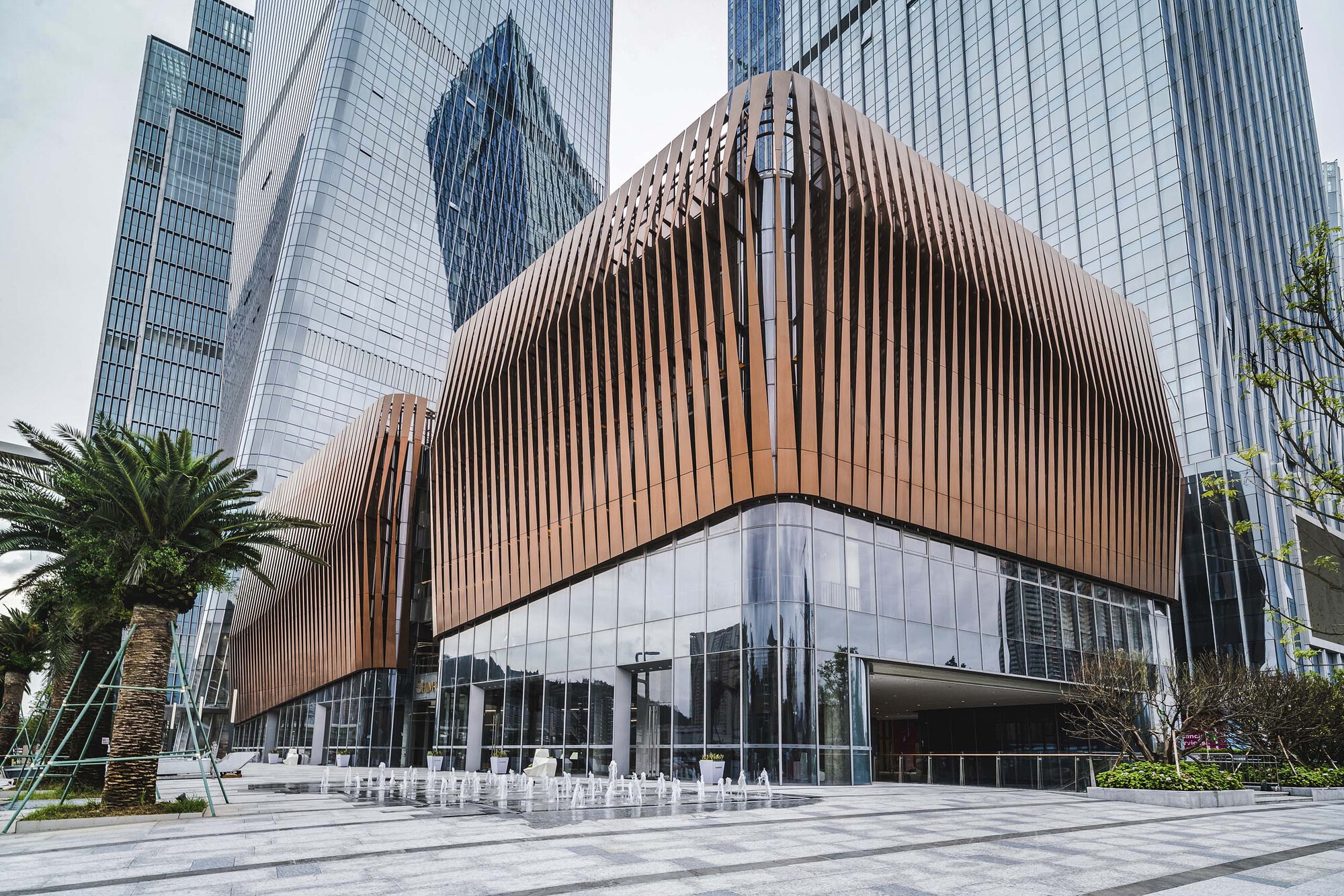 Curved copper-clad podium with vertical fins above a glazed base and plaza fountain with palm trees