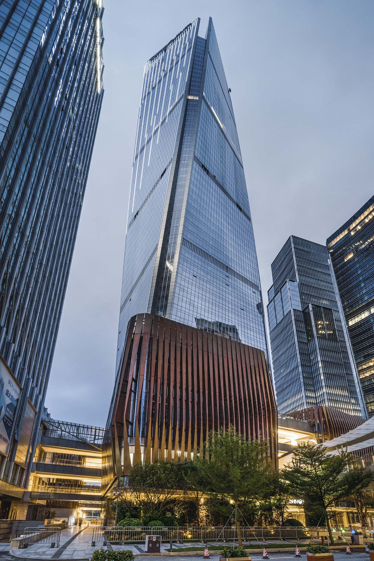 Upward view of the faceted glass tower with illuminated timber-clad base volume at twilight