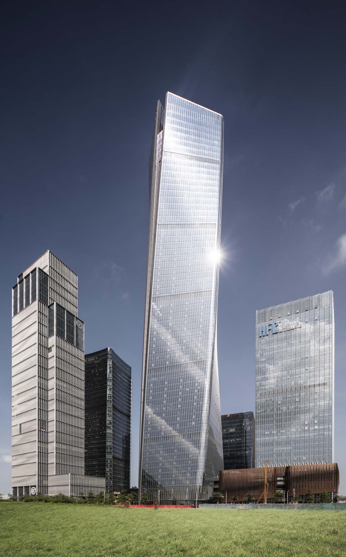 Twisted glass tower rising above surrounding buildings and a foreground lawn on a clear day