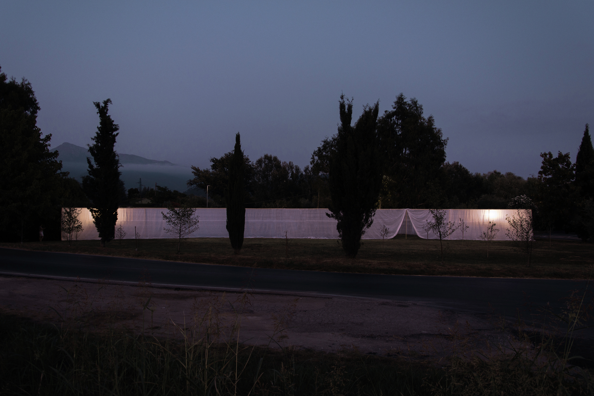 Illuminated fabric enclosure seen at dusk across a road with silhouetted cypress trees