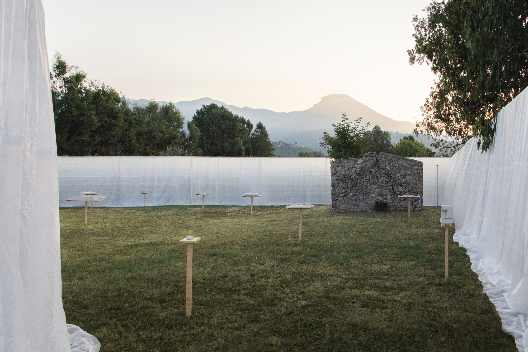 Grass courtyard framed by white fabric curtains with a stone wall and distant mountains at dusk