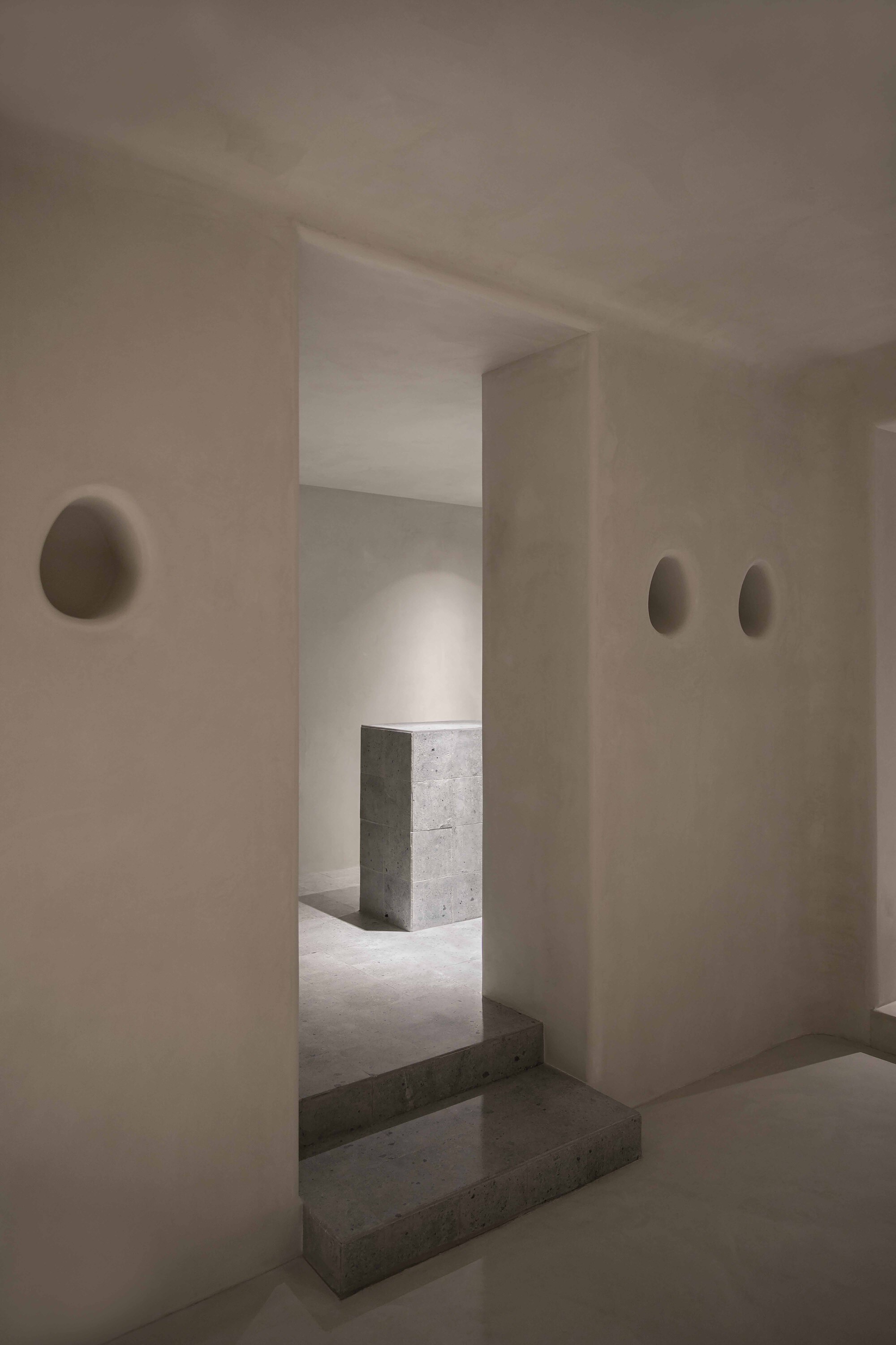Interior threshold with concrete steps connecting plastered rooms punctuated by oval wall sconces