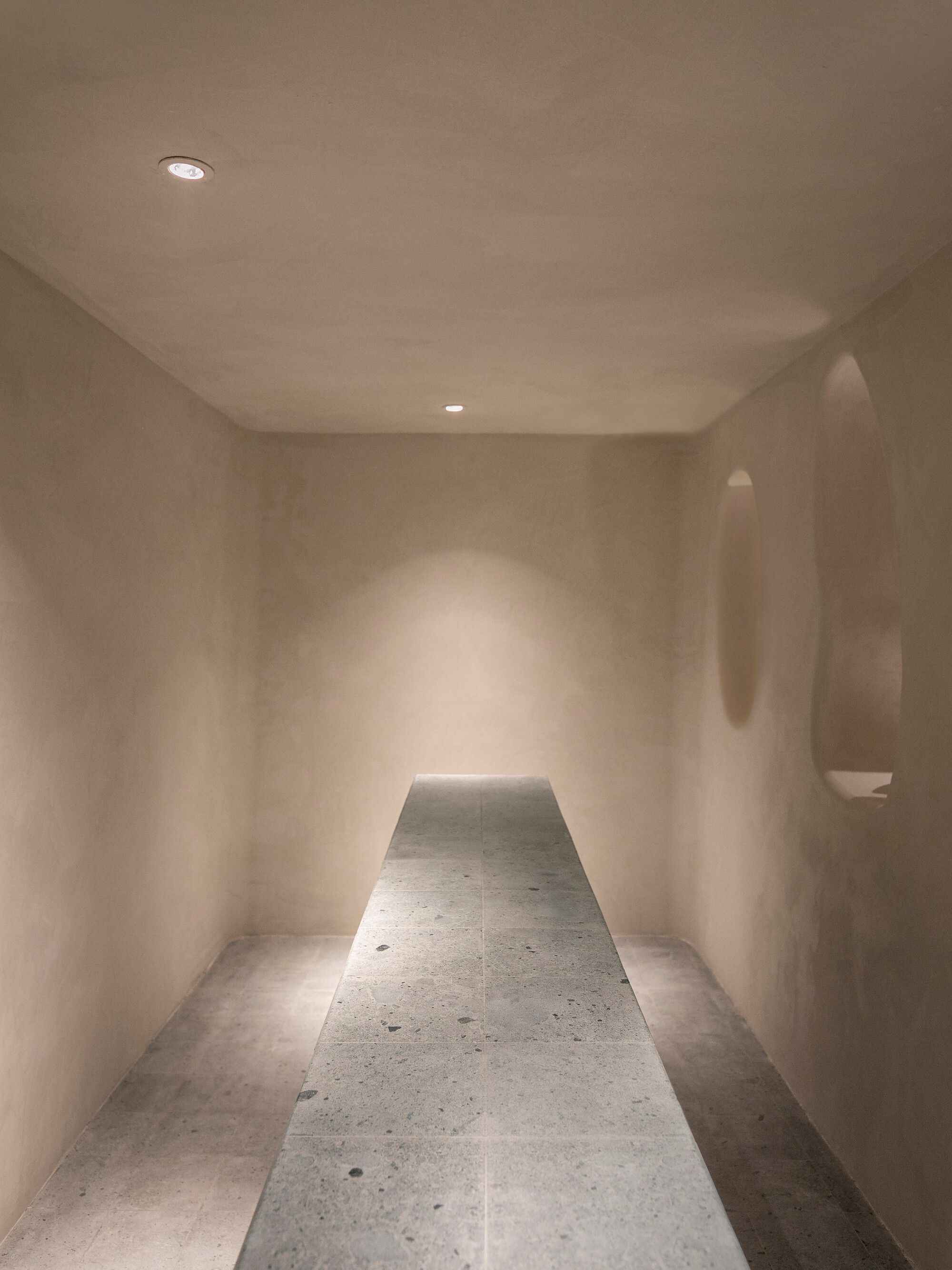 Narrow room with terrazzo floor panel and oval wall niches illuminated by recessed ceiling lights
