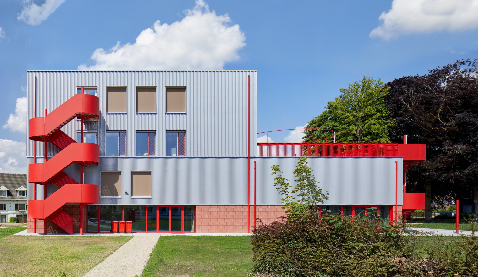 Facade showing corrugated metal panels, brick base, and red steel staircase beside young trees under cloudy skies