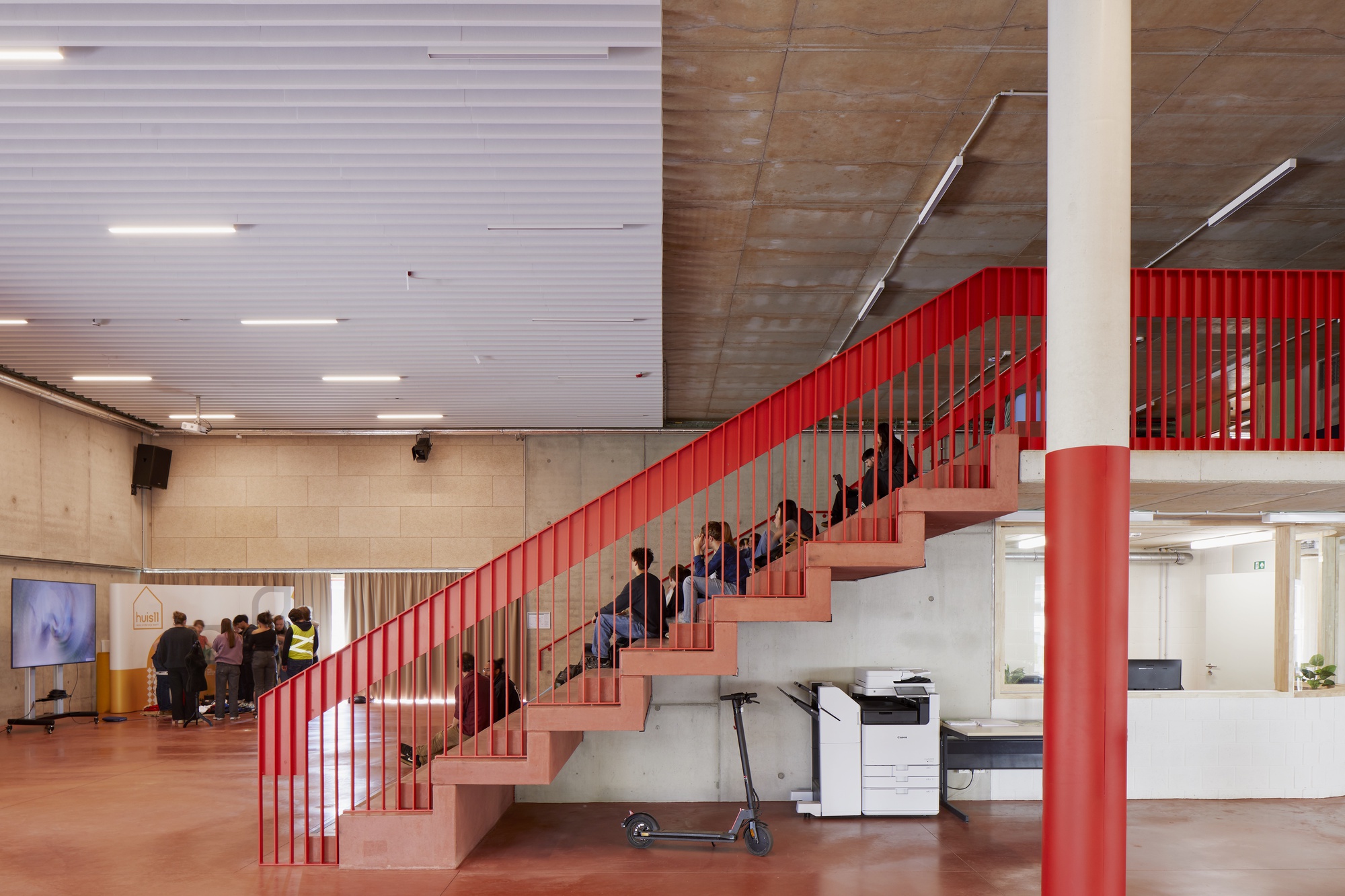 Interior atrium with red steel staircase, vertical balustrade, and concrete ceiling with fluorescent lighting above a gathering