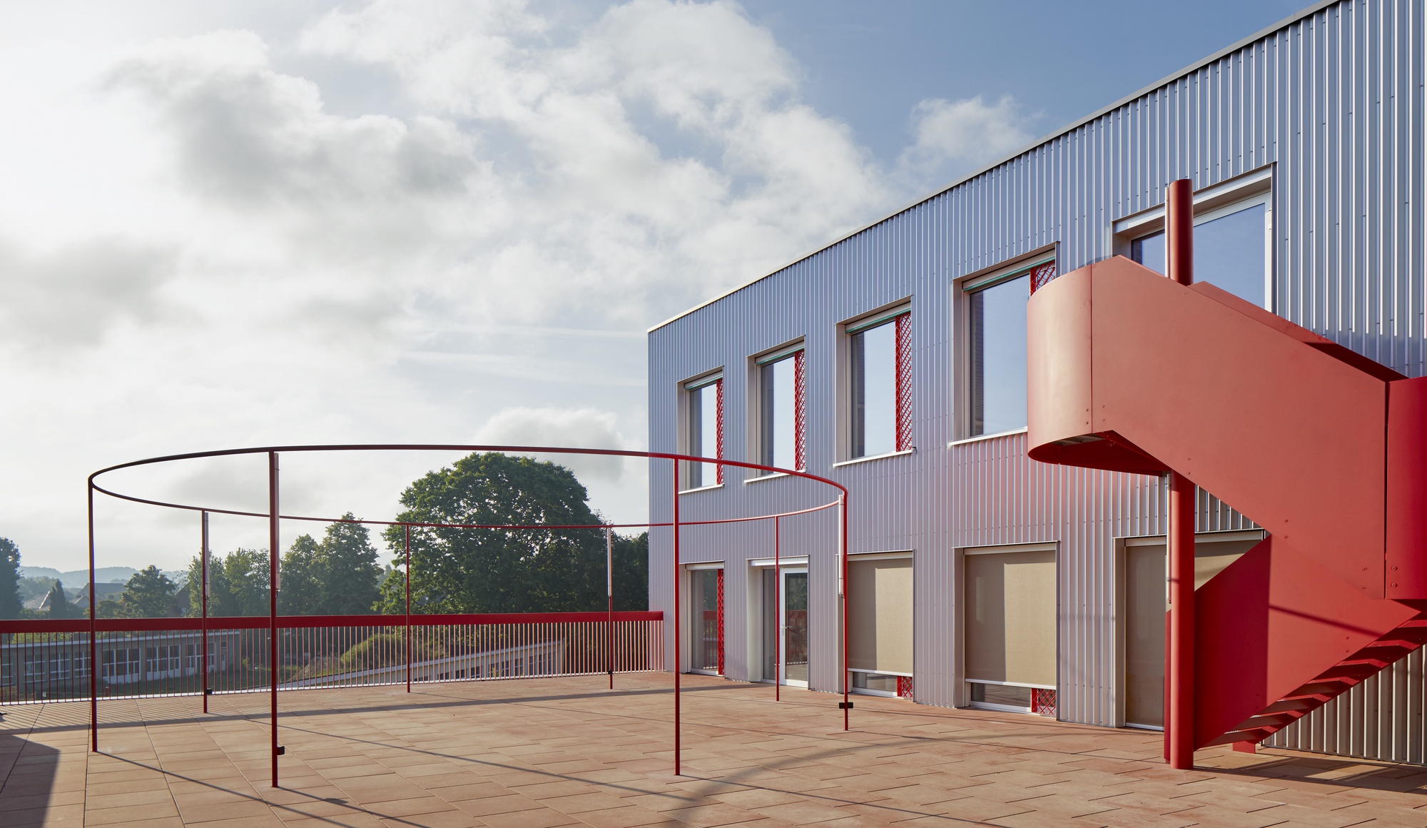 Rooftop terrace with red steel railing, play equipment, and external staircase beside corrugated metal facade