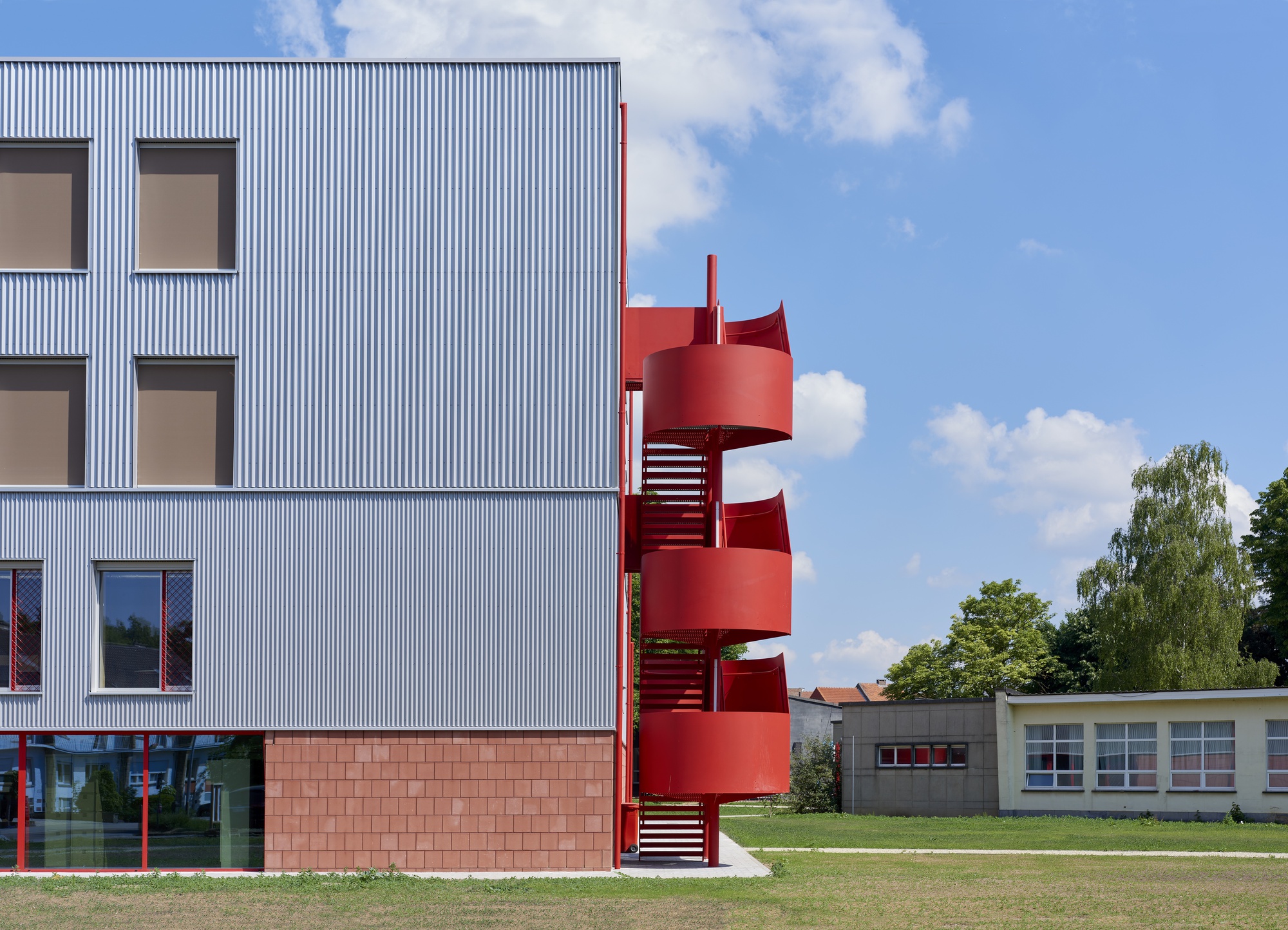 Close view of the red cylindrical staircase wrapping the corrugated metal facade under a blue summer sky