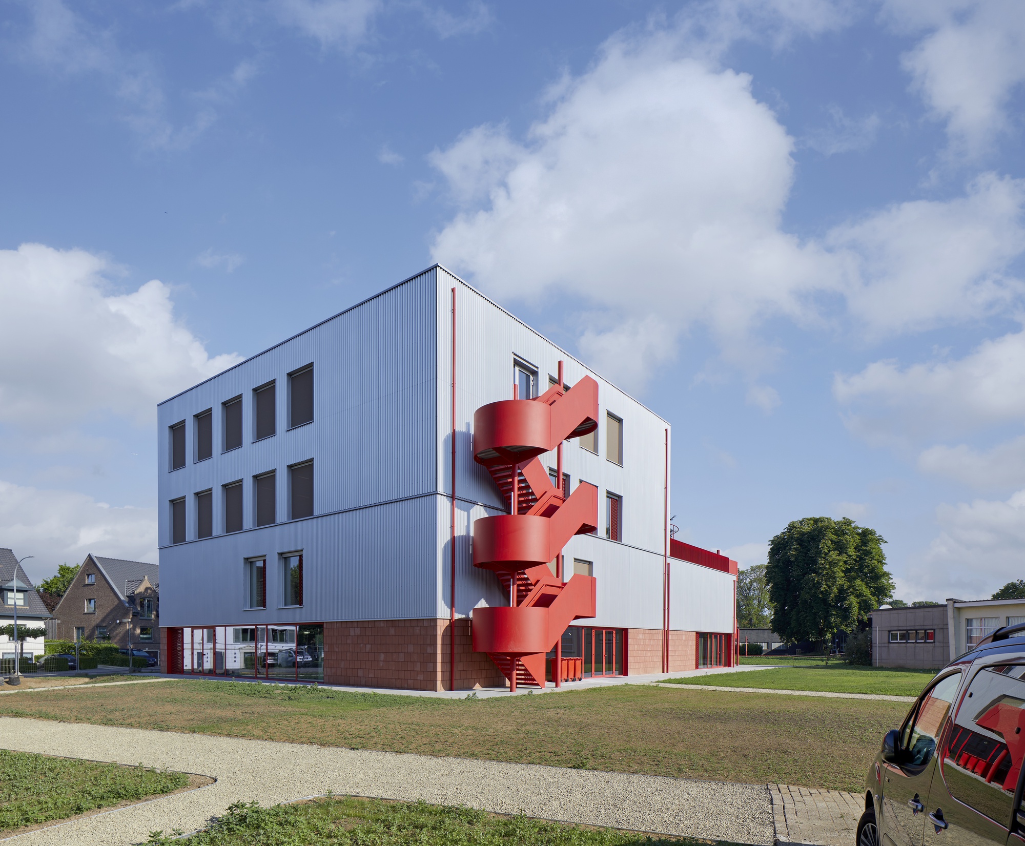 Street view showing the corrugated metal facade with a sculptural red stair tower against a partly cloudy sky
