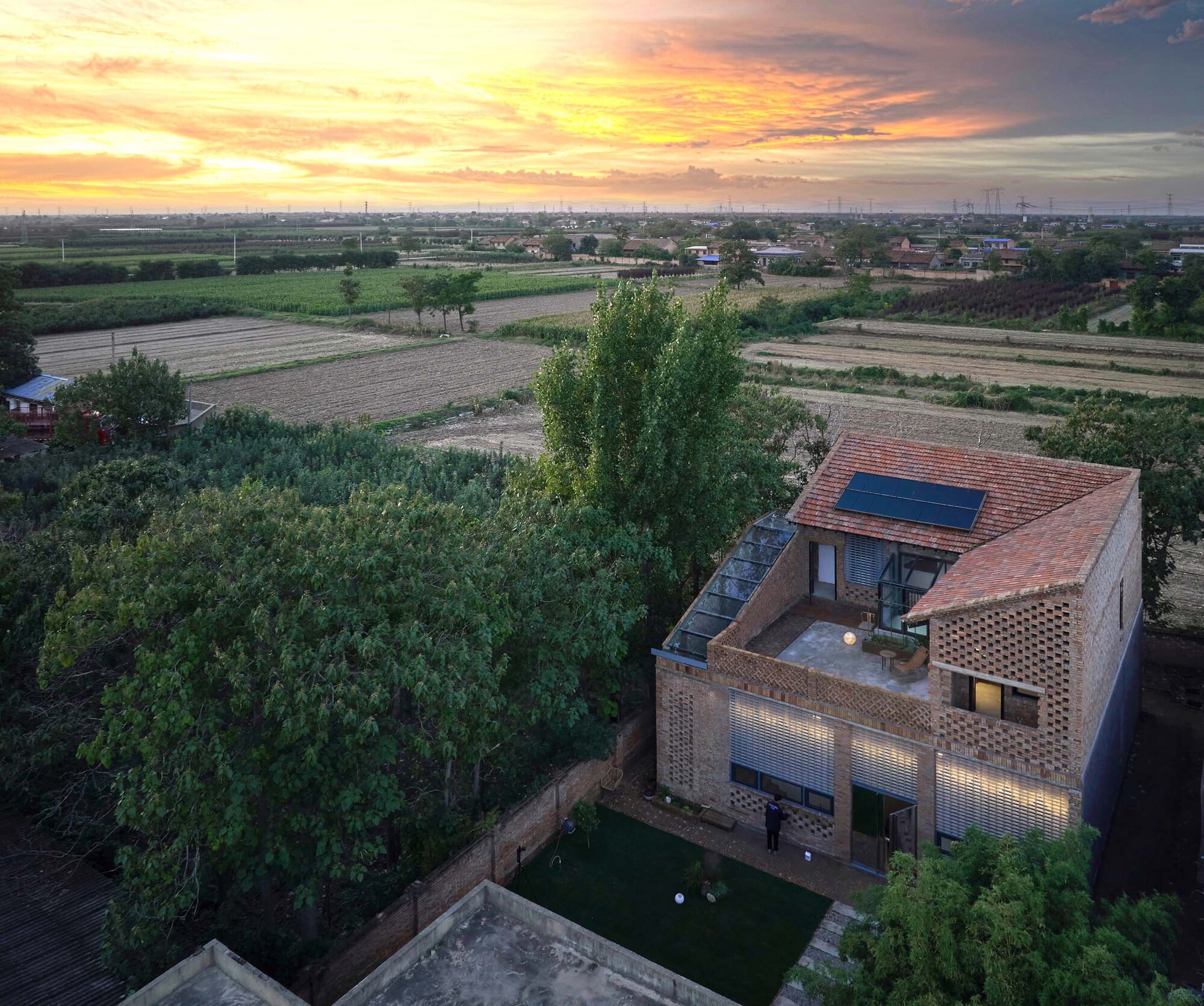 Aerial view of the brick dwelling with illuminated glass block facade at sunset above agricultural fields