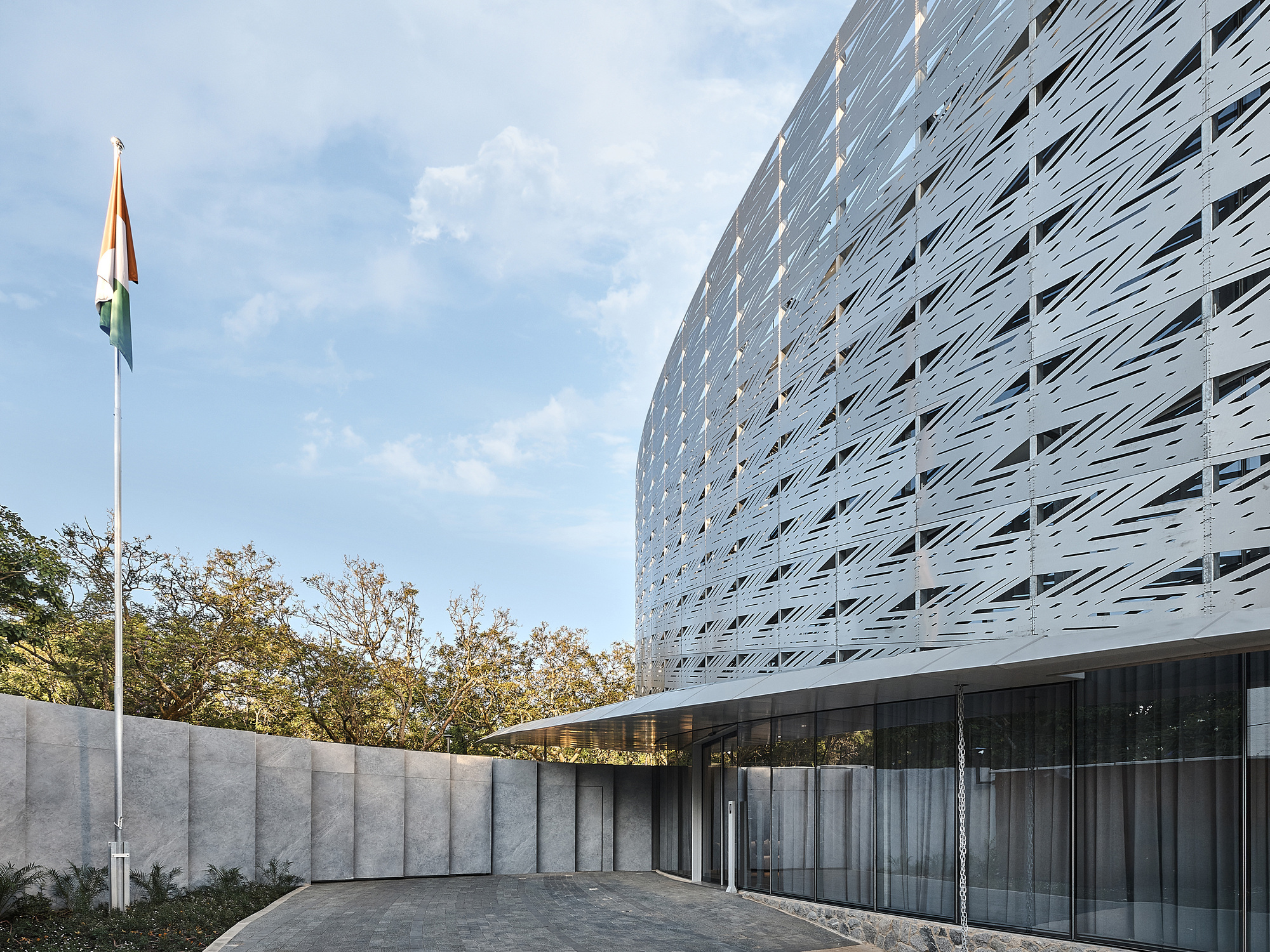 Ground-level view of the perforated metal facade meeting a concrete wall and glazed entrance below afternoon clouds