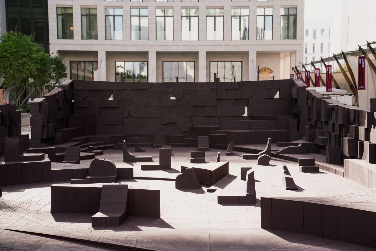 Public plaza with dark brown sculptural seating elements and stepped amphitheater wall beneath surrounding buildings