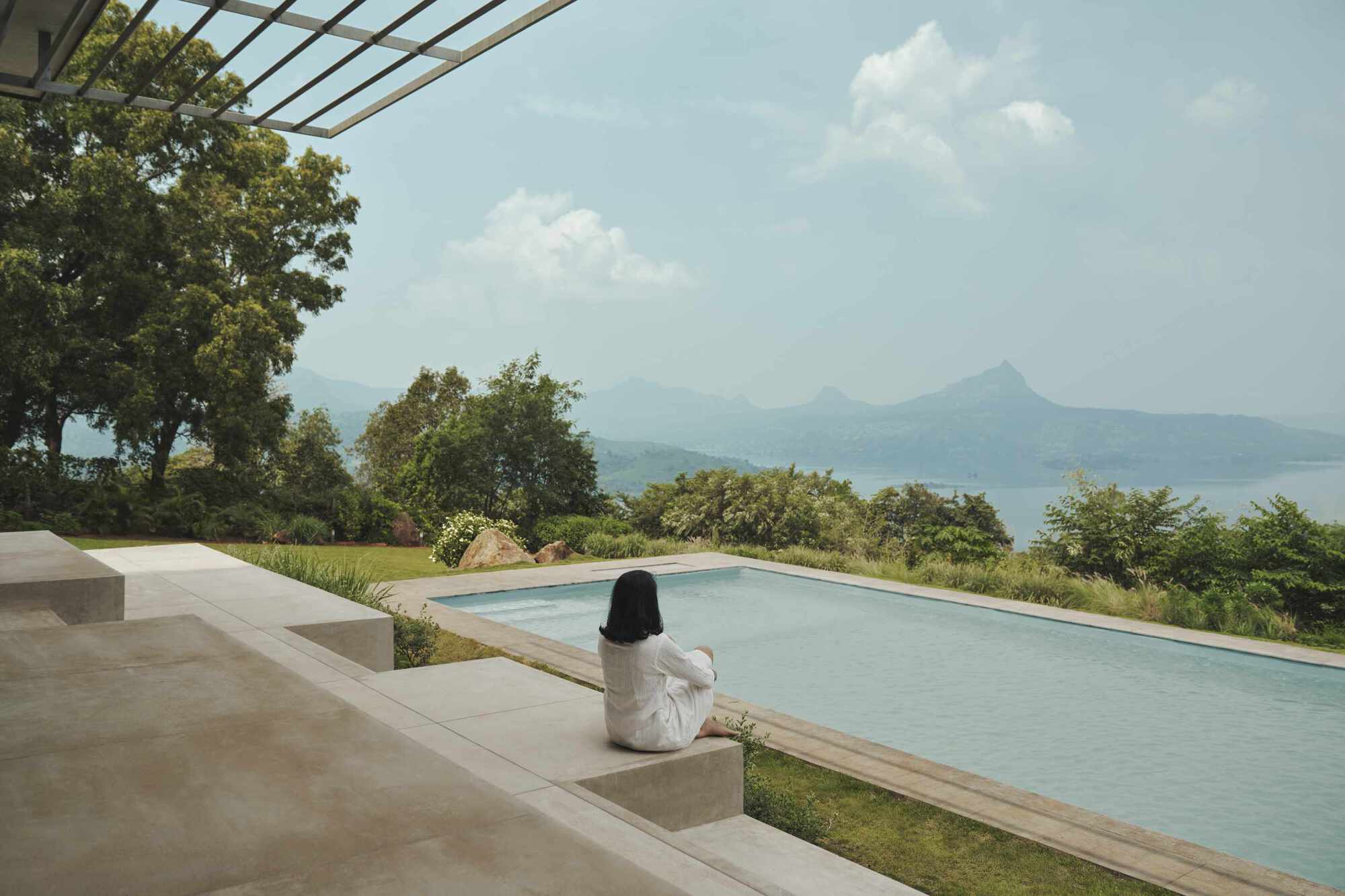 Infinity pool terrace with concrete seating ledge where a person sits overlooking mountains and lake