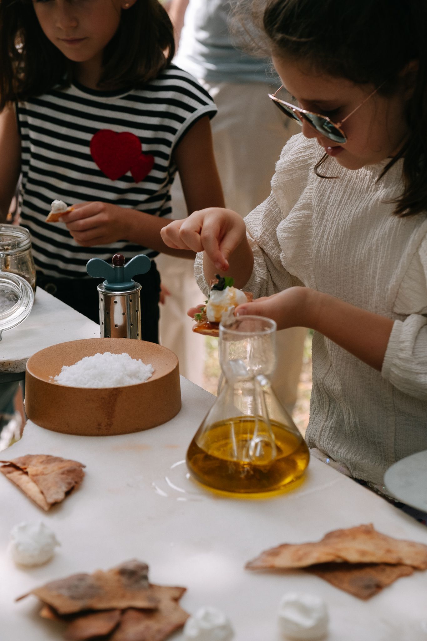 Two children preparing food at a white table with olive oil, salt, and flatbread