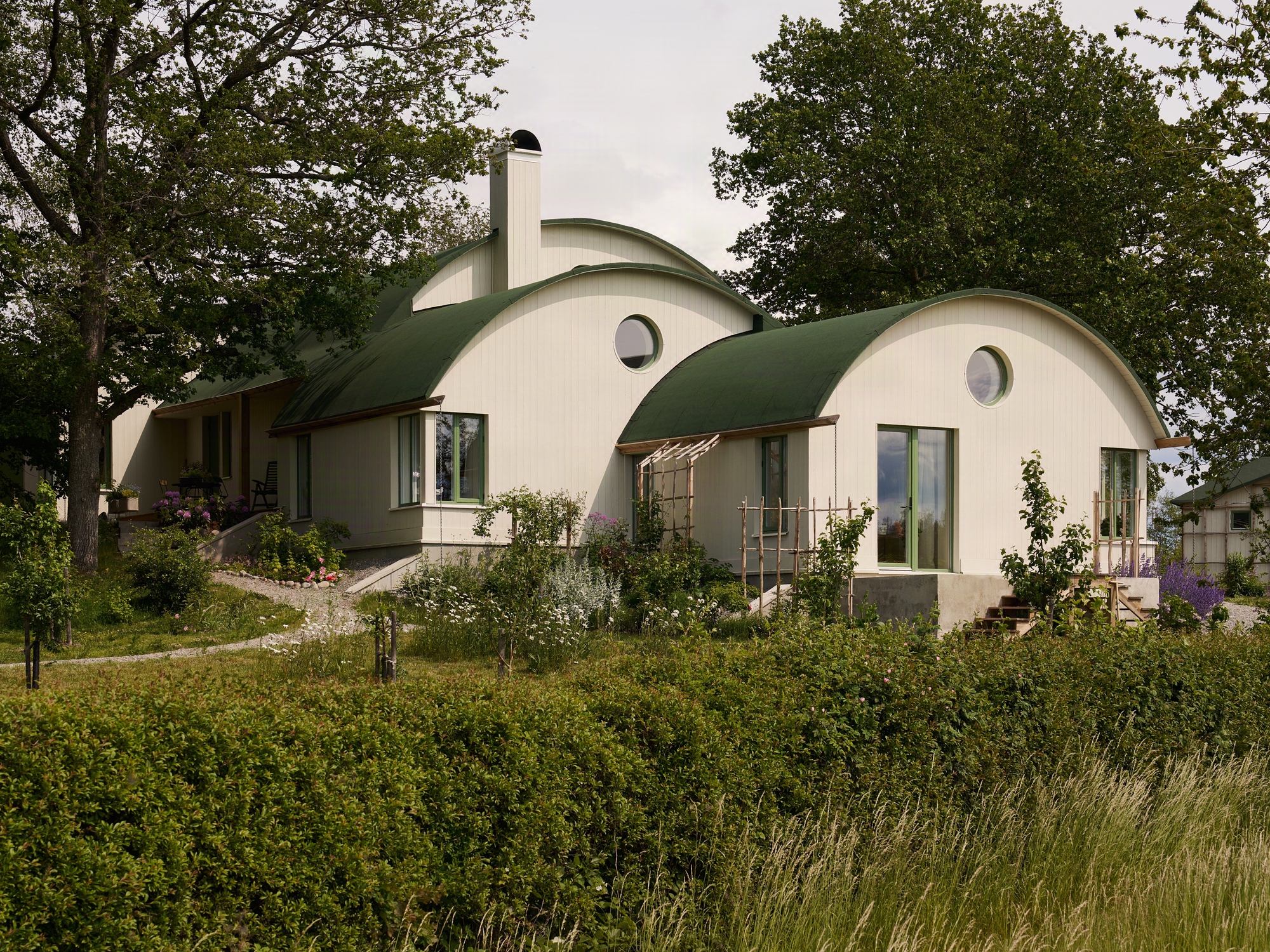 Garden elevation showing the three vaulted volumes with planted beds and a timber pergola in afternoon light
