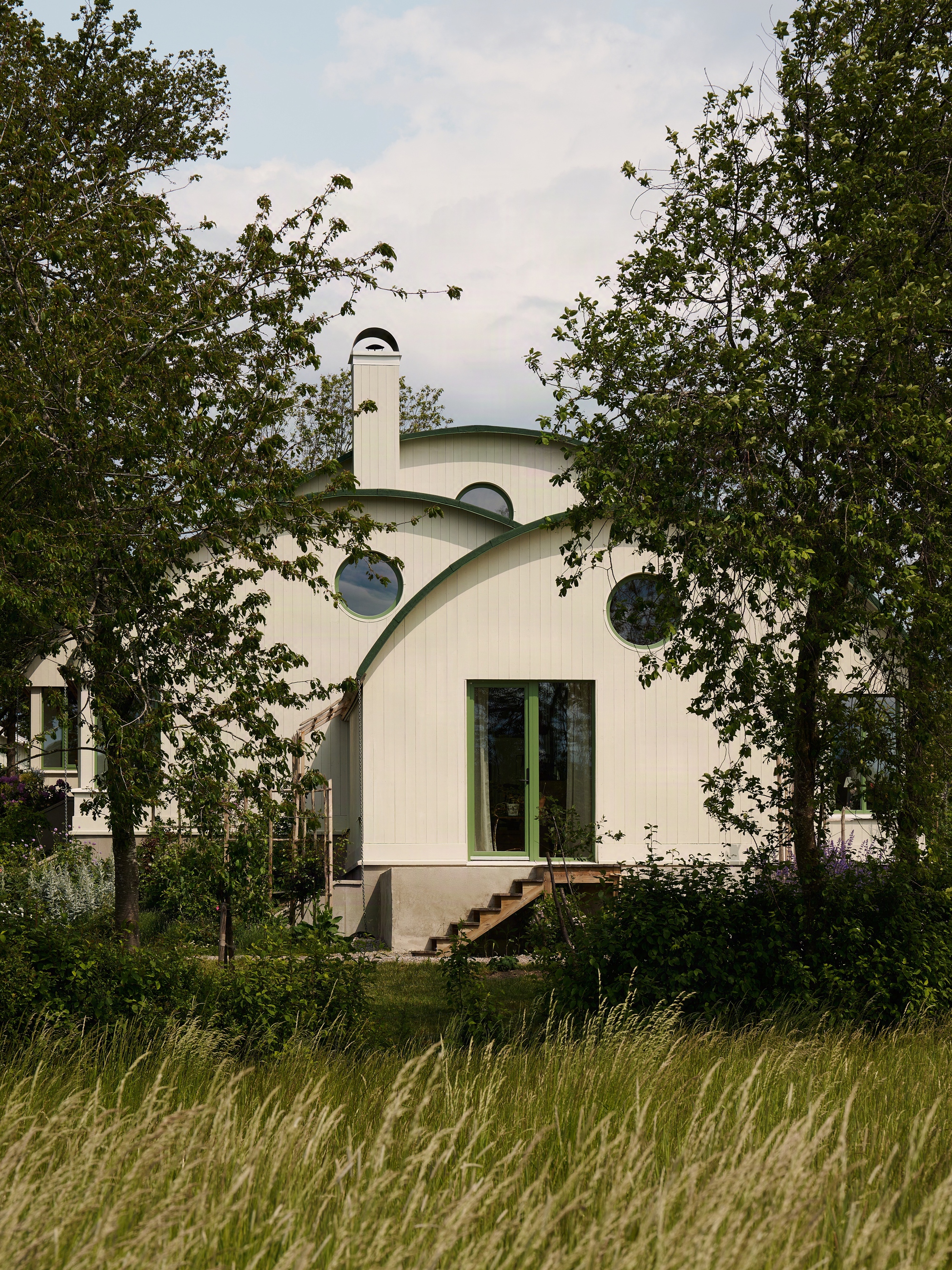 White vertical wood siding and curved green roof with round windows framed by tall trees