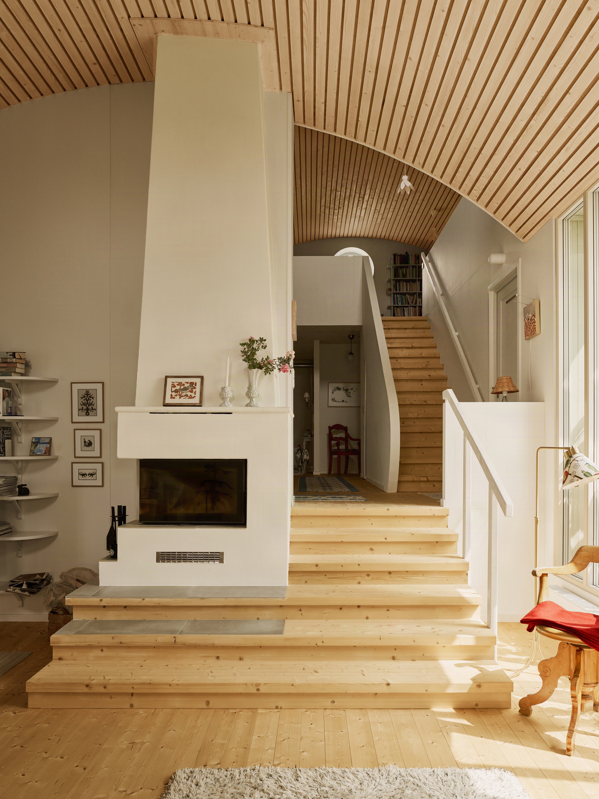 Split-level interior with pale timber stairs rising past a fireplace under curved wood slatted vaulting