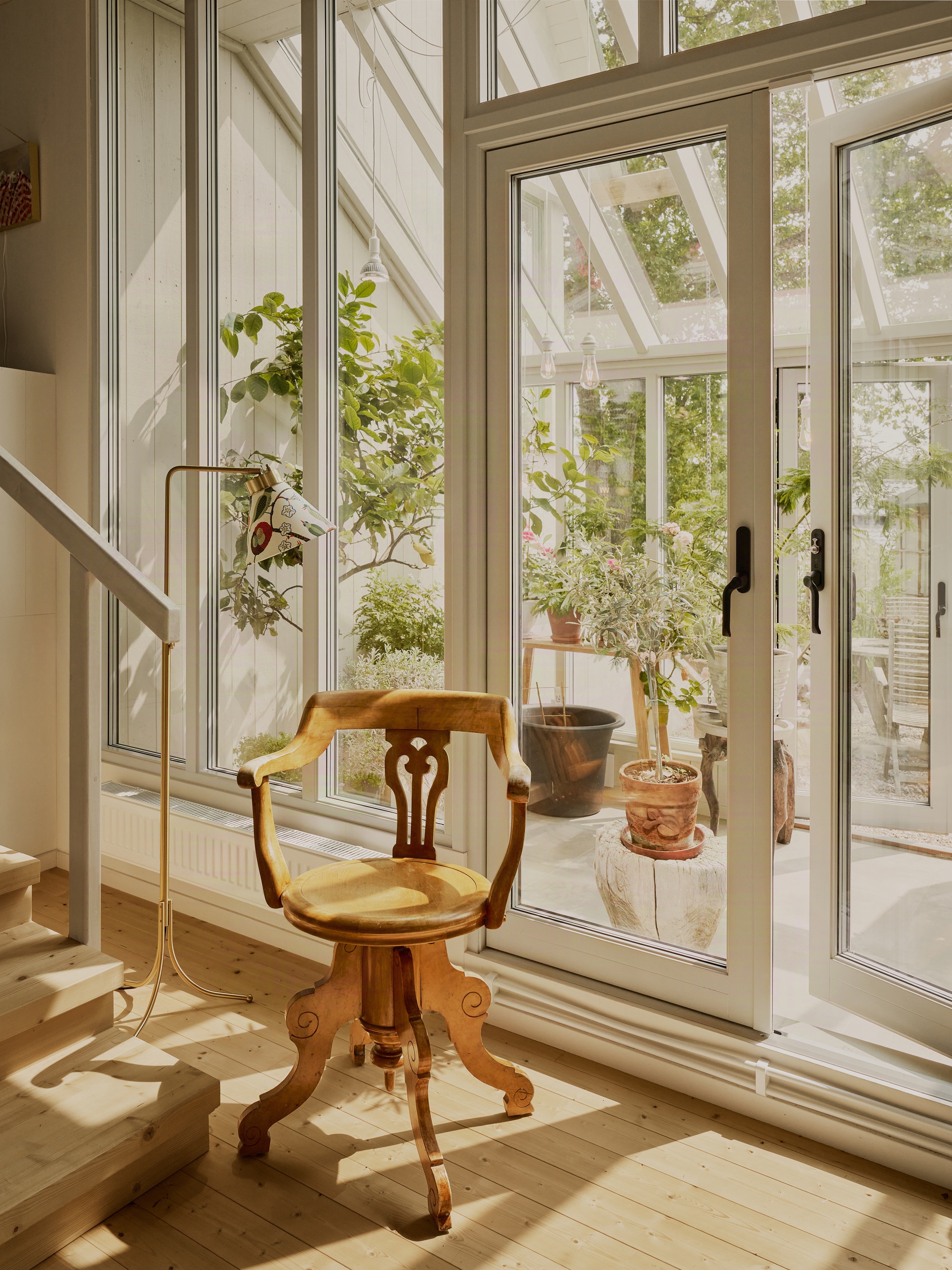Glazed corner with full-height white-framed doors opening to a greenhouse with potted plants in sunlight