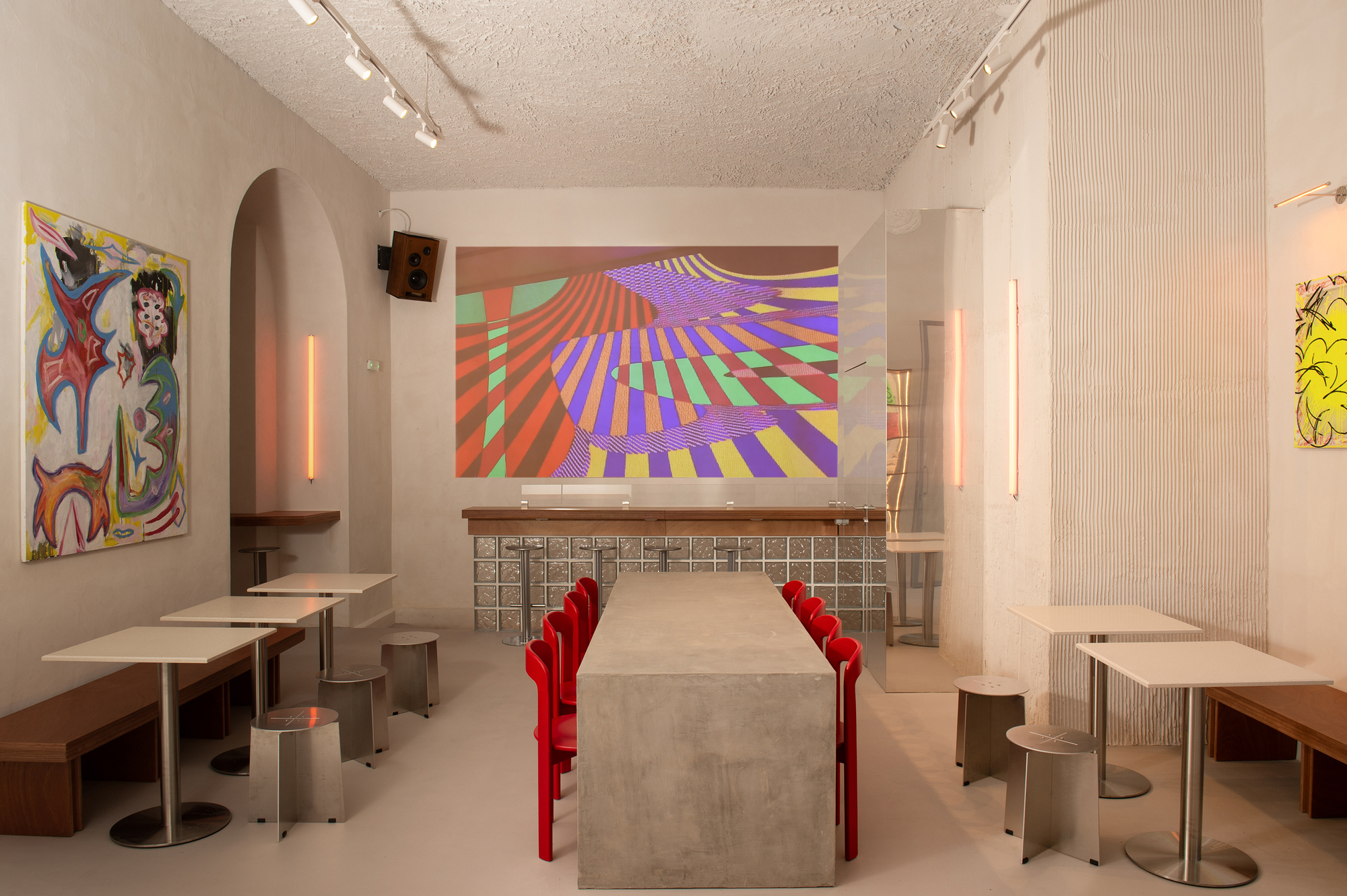 Interior dining area with concrete tables, red chairs, white mosaic tile bar and projected colorful geometric artwork