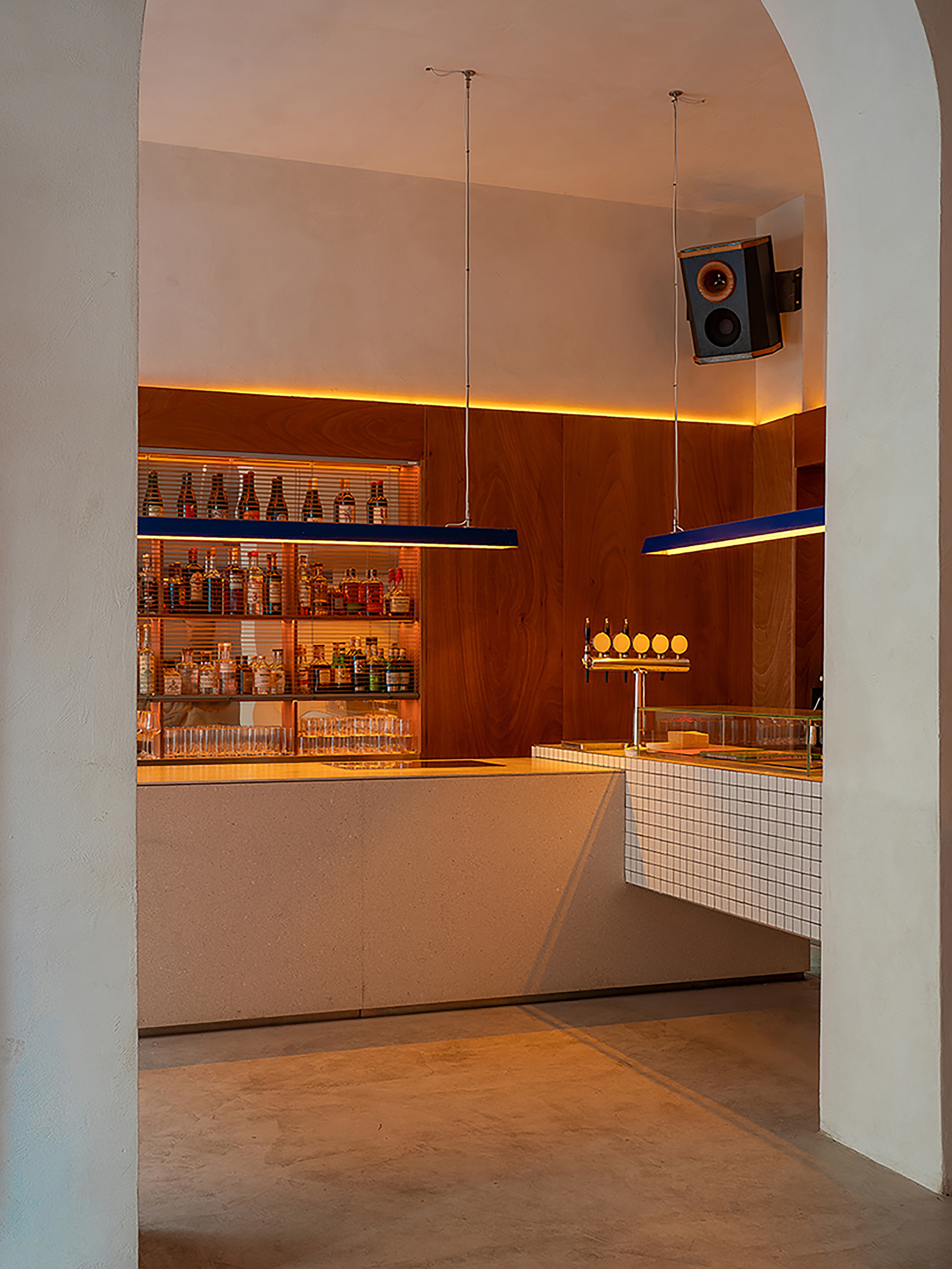 Bar counter with white mosaic tile front, timber paneling, backlit bottle shelves and suspended speakers