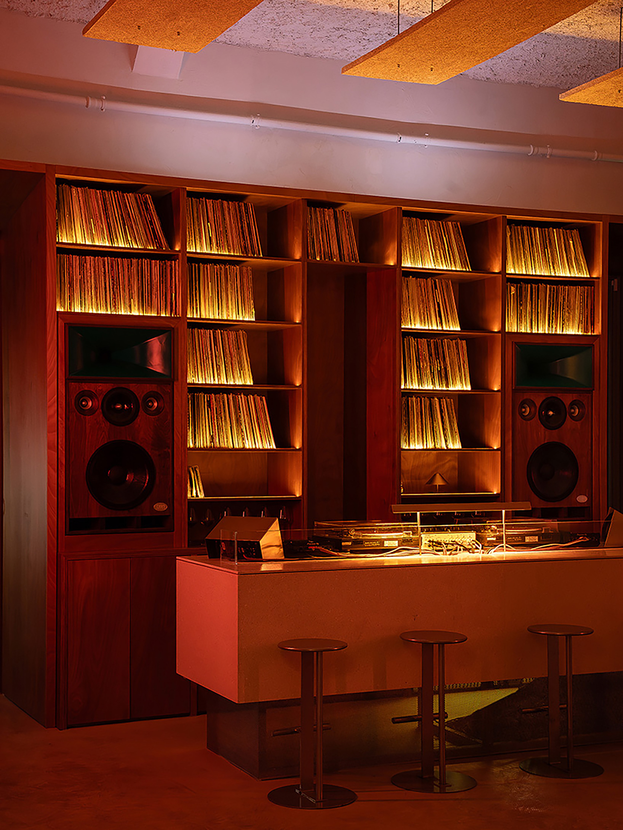 Listening bar with vinyl shelves and integrated speakers beneath acoustic ceiling panels under warm lighting