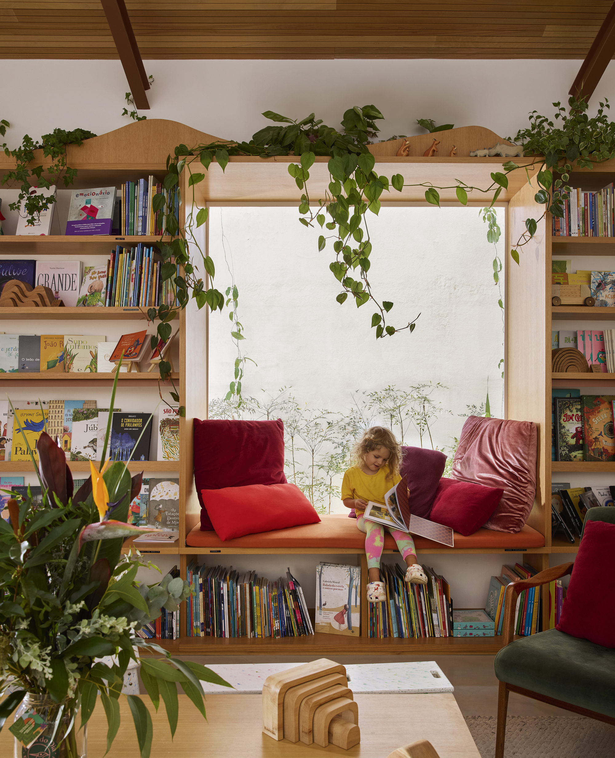 Built-in window seat with plywood bookshelves and cascading potted vines under exposed timber beams with child reading