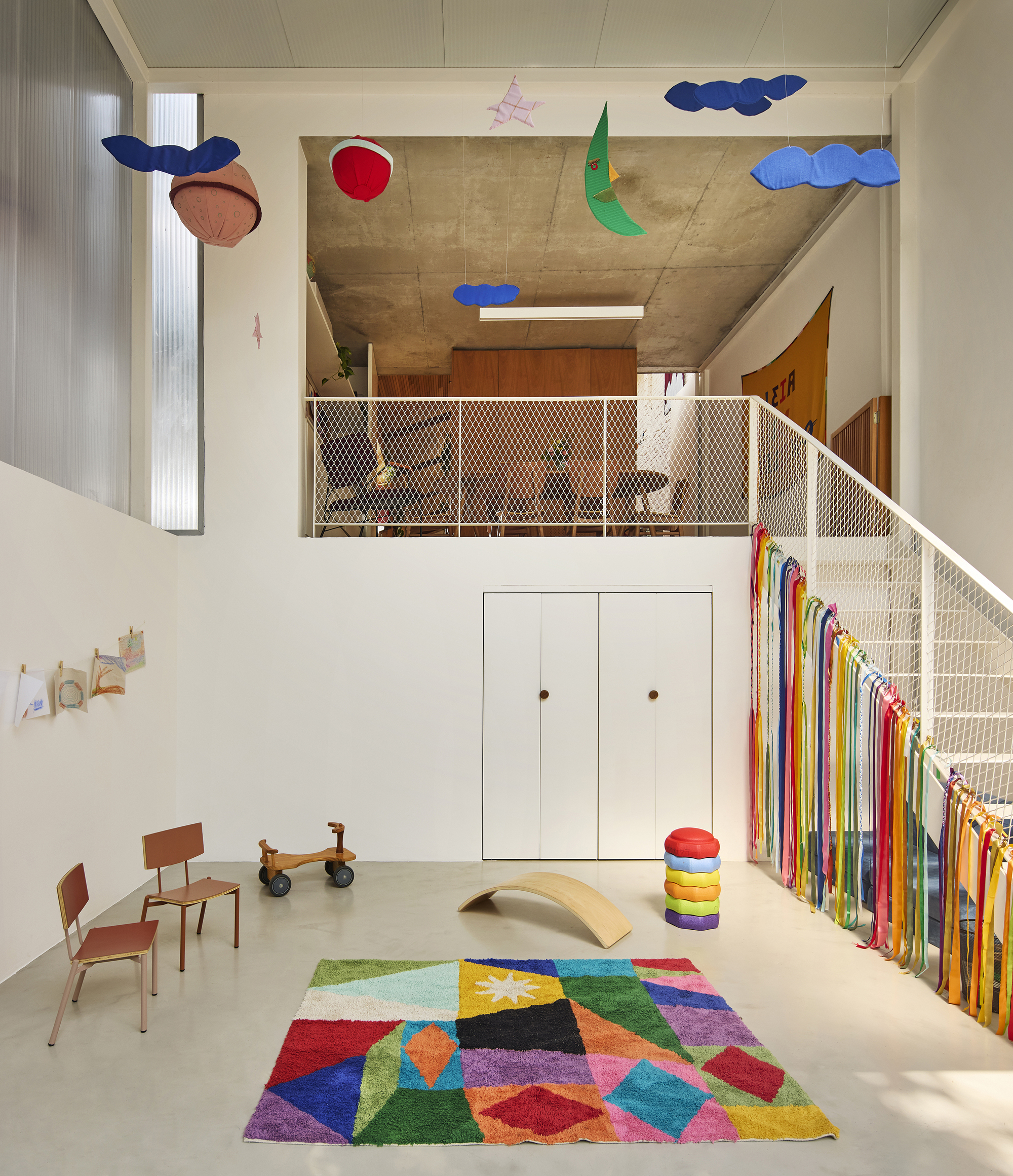 Double-height playroom with mesh-railed mezzanine, exposed concrete soffit, and suspended fabric mobiles above multicolored geometric rug