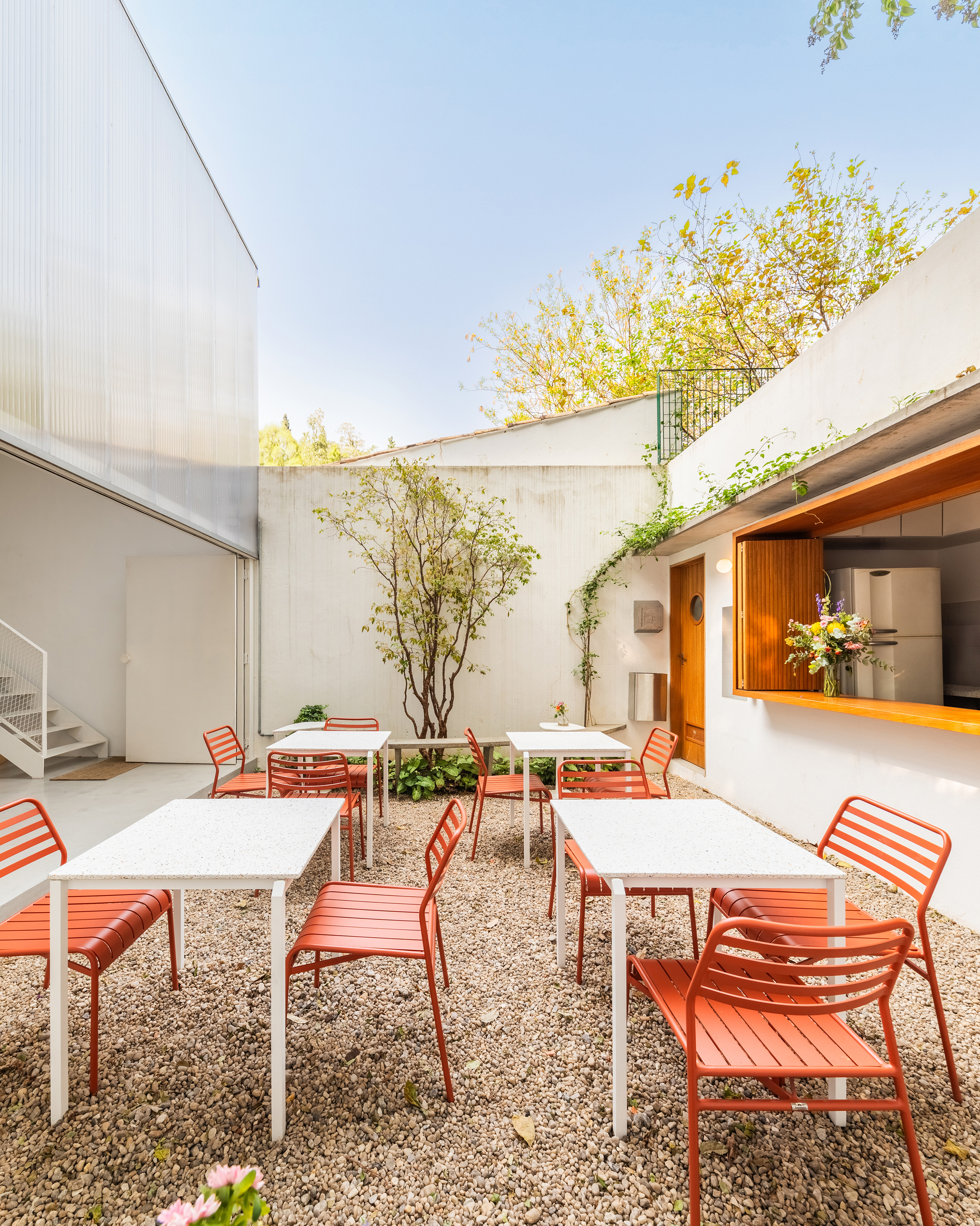 Gravel courtyard with red metal seating and white tables framed by concrete walls and deciduous trees