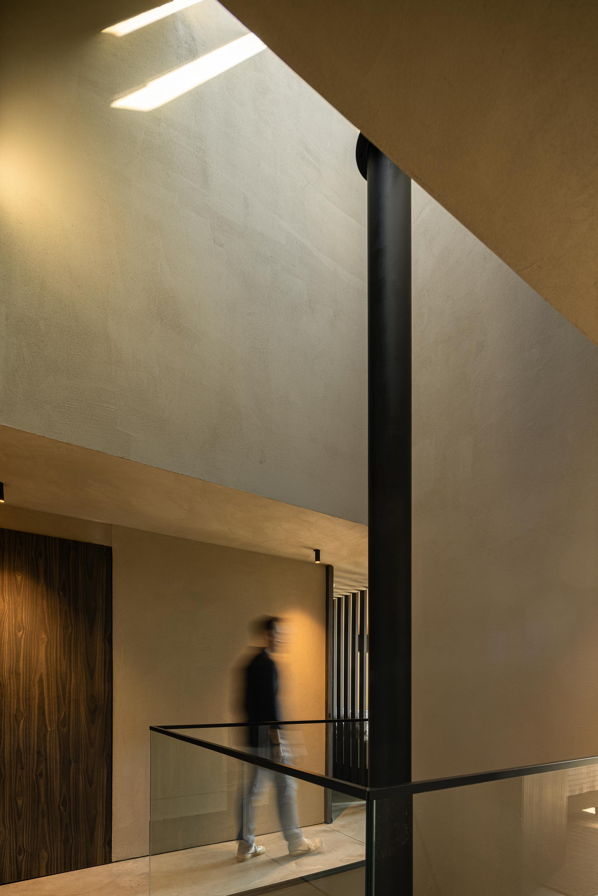 Double height hallway with angled skylight illuminating plaster walls and a figure crossing glass balcony