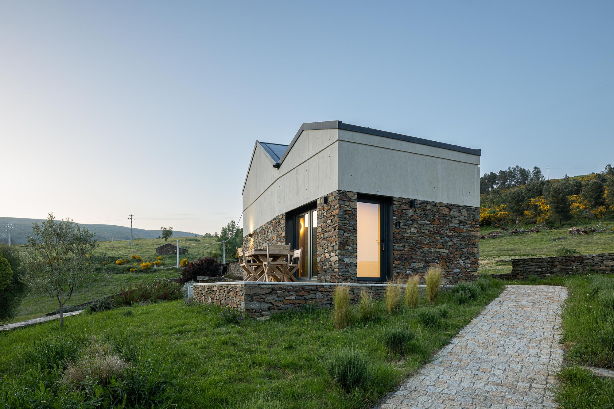 Hillside residence with stone plinth and concrete upper volumes at twilight with cobbled path